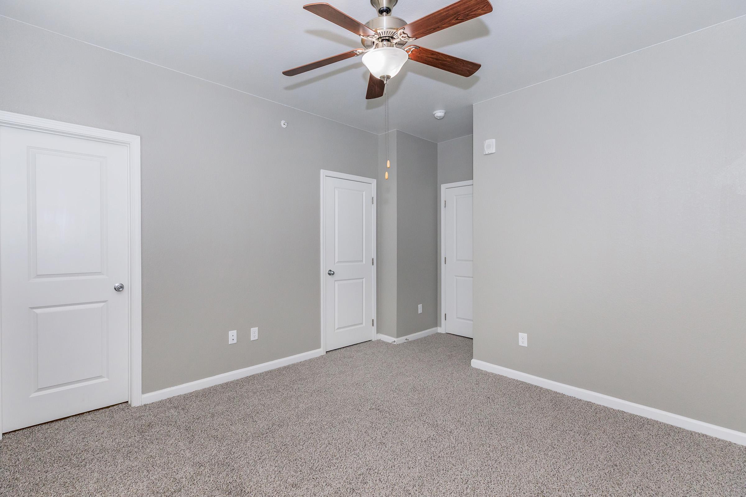A spacious, empty room featuring light gray walls and a ceiling fan with wooden blades. There are two closed white doors on adjacent walls, and the floor is covered with light-colored carpet. The room is well-lit, enhancing its openness and neutrality, ideal for various interior design options.