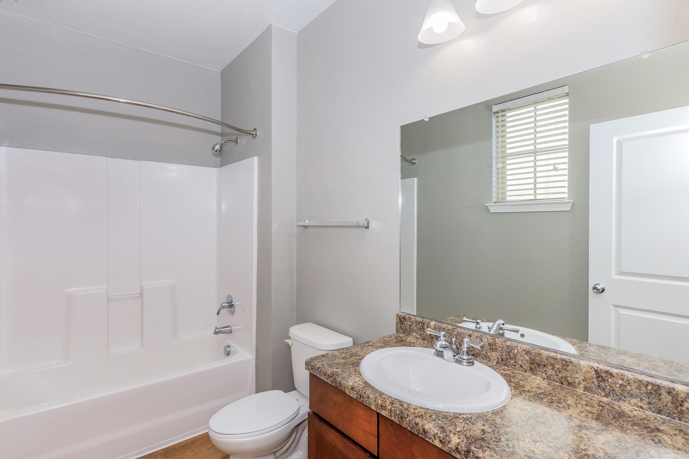 A modern bathroom featuring a white bathtub with a shower, a vanity with a sink and brown cabinets, a toilet, and a large mirror above the sink. The walls are painted a light grey, and natural light streams in through a window with white blinds.