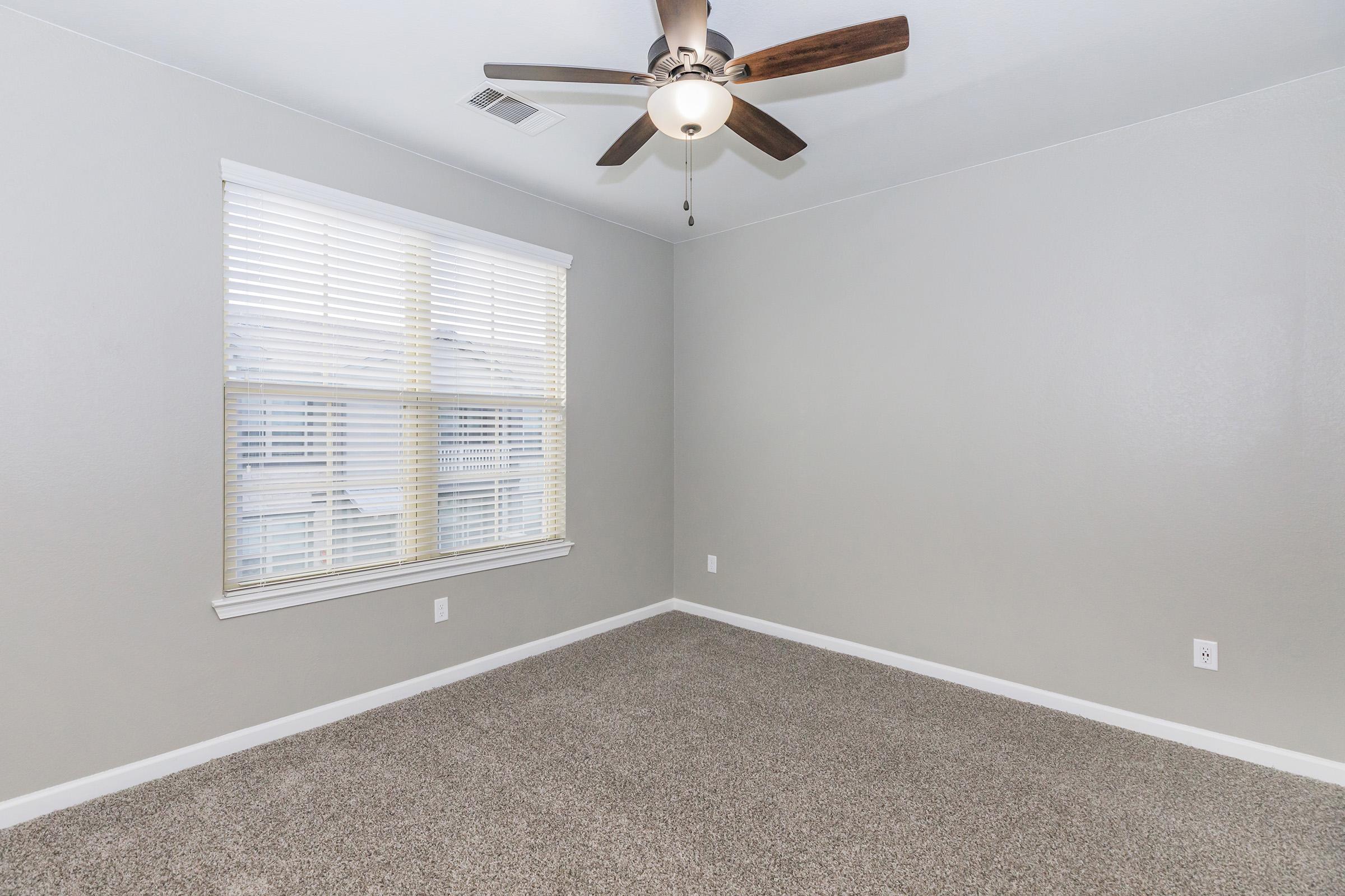 A vacant room with light gray walls and carpeted flooring. It features a ceiling fan with wooden blades and a window with white blinds that allows natural light to enter. The room has minimal decoration and no furniture, creating a spacious and airy atmosphere.