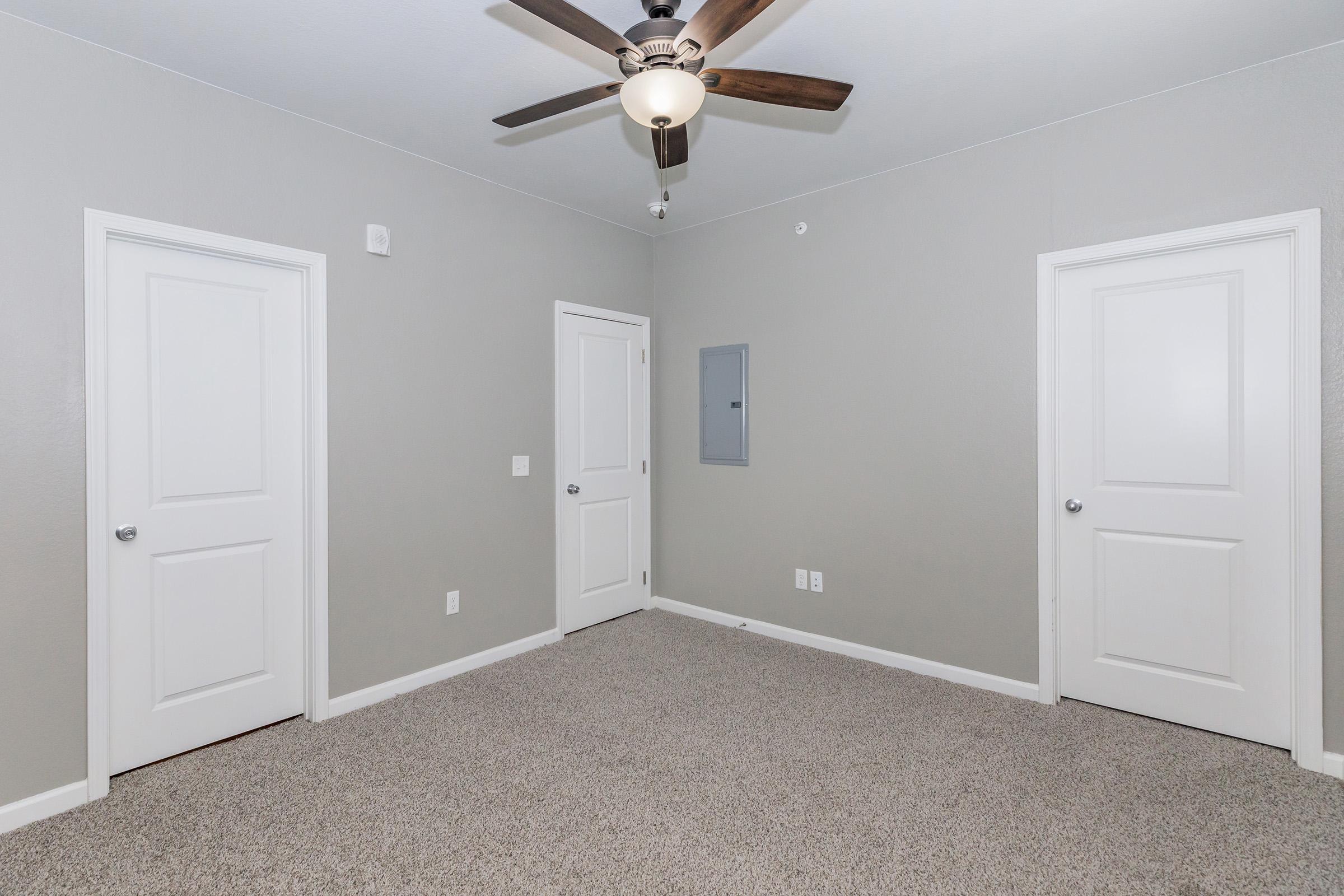 Empty room with light gray walls and carpeted floor. Two white doors are visible on opposite walls. A panel on the wall and a ceiling fan are also present. The room is well-lit and has a minimalistic design, creating an open and spacious feel.
