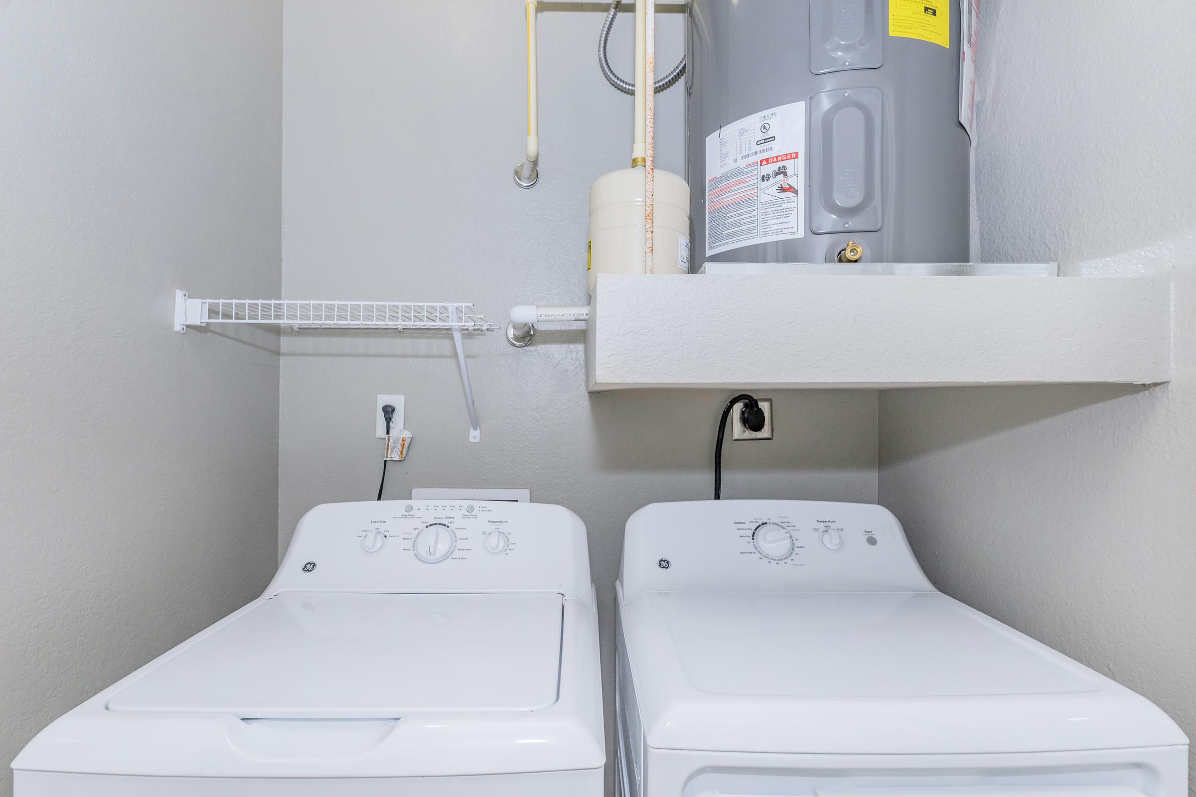 A laundry room featuring two white washing machines side by side, with a shelf above them. A water heater is mounted on the wall above the shelf, and there are exposed pipes. The walls are painted a light gray color, creating a clean and organized appearance.
