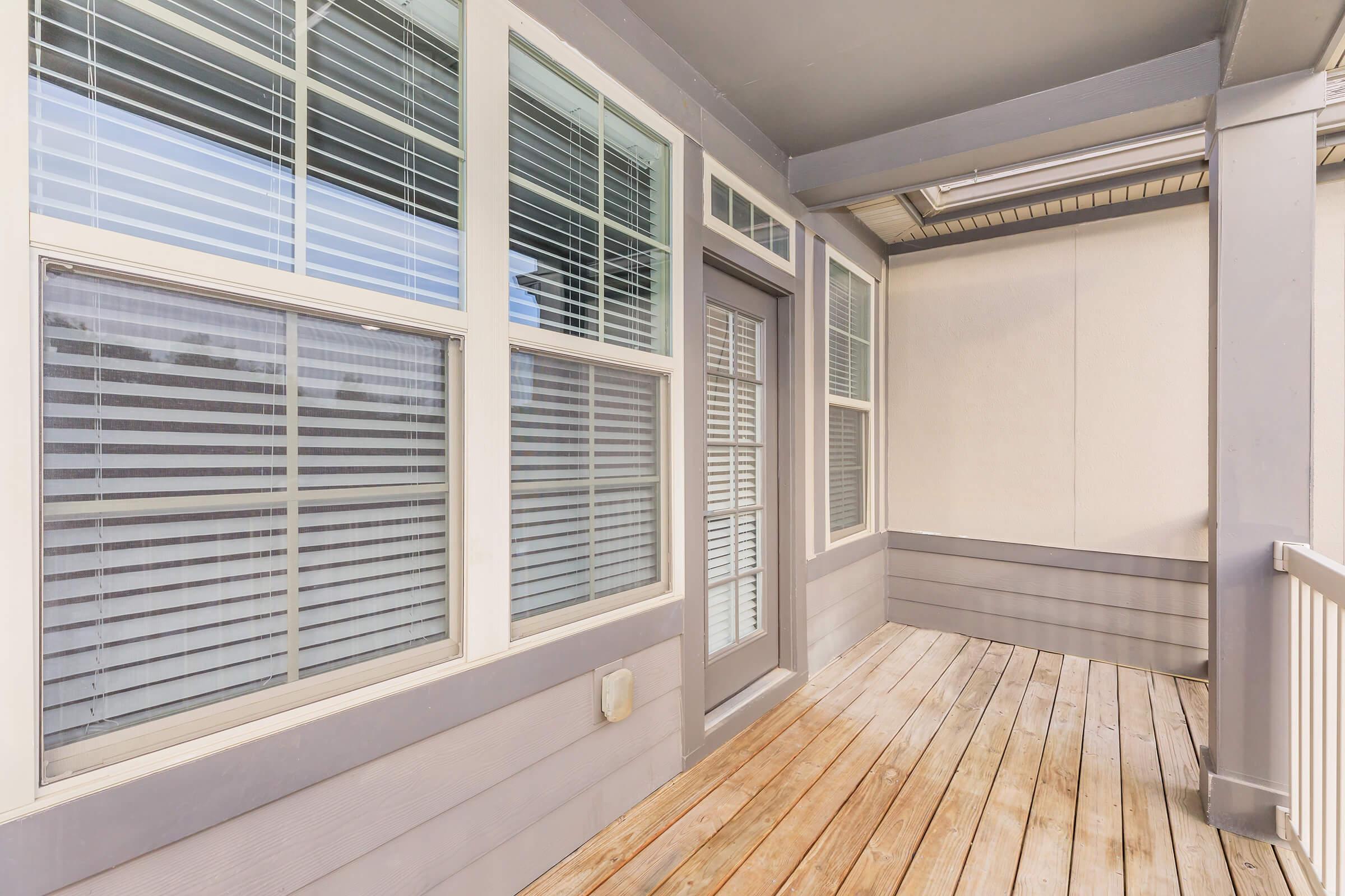 A well-lit outdoor patio space featuring large windows with blinds on the walls, a door leading outside, and wooden flooring. The walls are painted in a light gray color, creating a modern and inviting atmosphere. Ideal for relaxing or entertaining guests.
