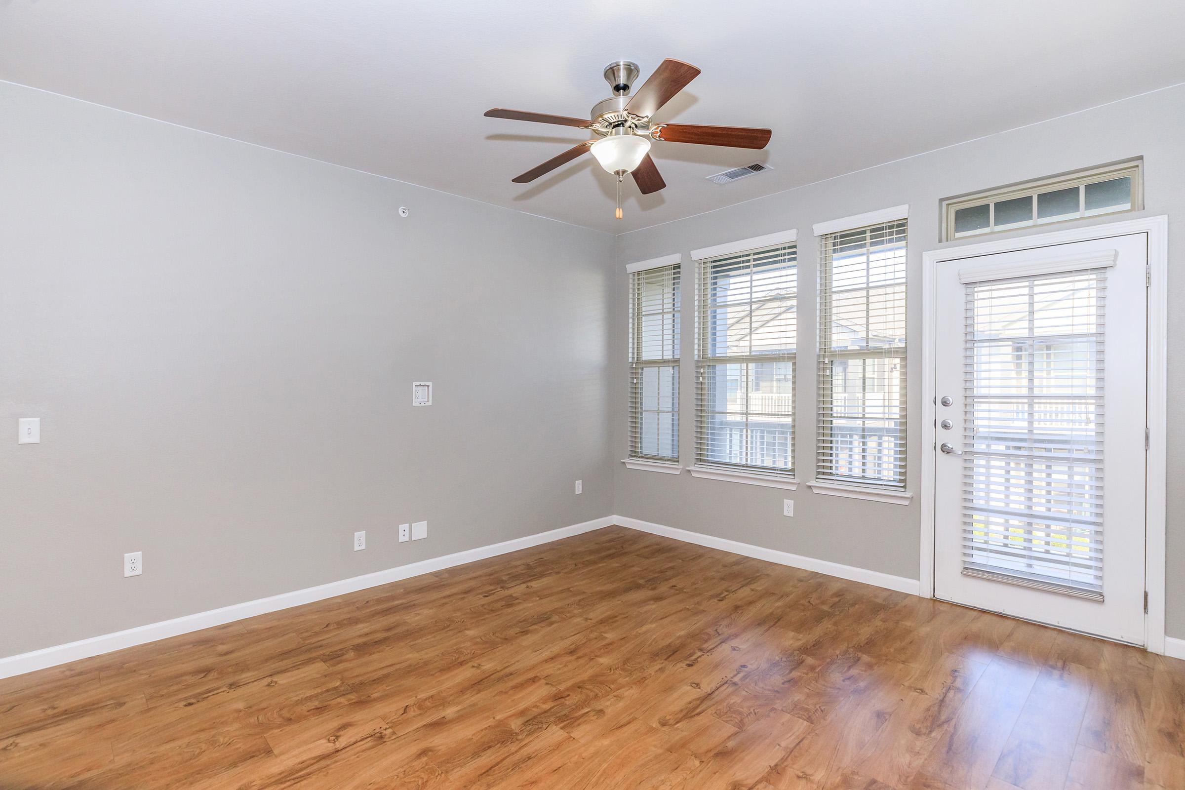 A spacious room featuring a ceiling fan, large windows with blinds allowing natural light, and a door leading outside. The hardwood floor adds warmth to the modern, neutral-toned walls, creating a welcoming atmosphere.