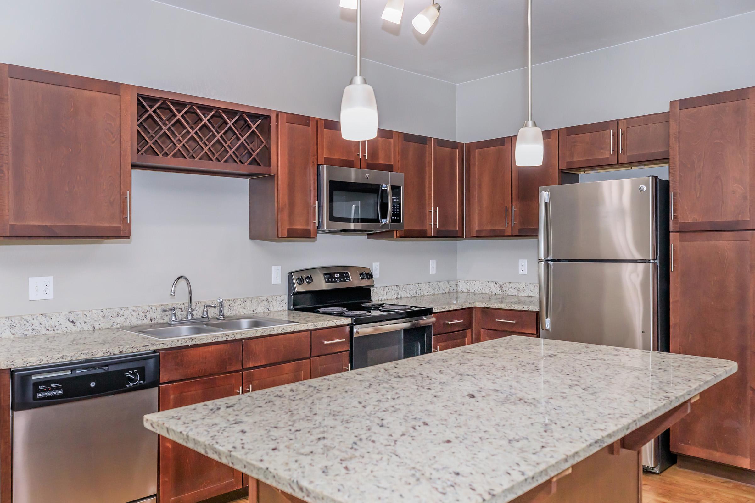 Modern kitchen featuring wooden cabinets, stainless steel appliances, and granite countertops. Includes an island with seating, a double sink, and a wine rack. Bright lighting from pendant lights enhances the clean and spacious layout, with a focus on functionality and style.