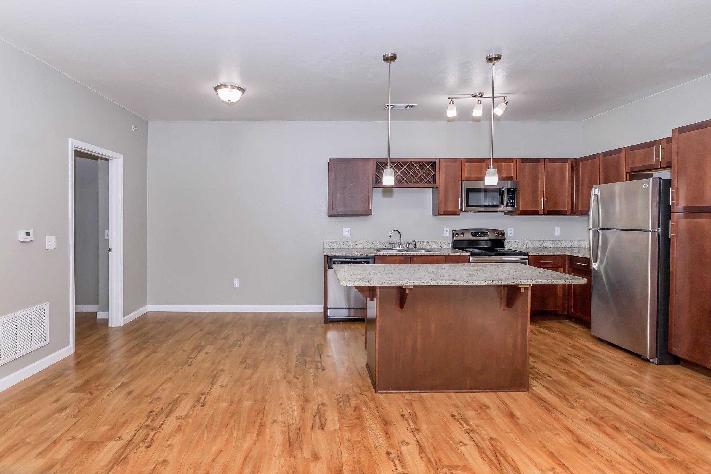 Modern kitchen featuring wooden cabinetry, granite countertops, and stainless steel appliances. The layout includes an island with seating space. Natural light fills the room, which has a neutral wall color and a spacious, open design with wood flooring. A door leads to another room on the left.