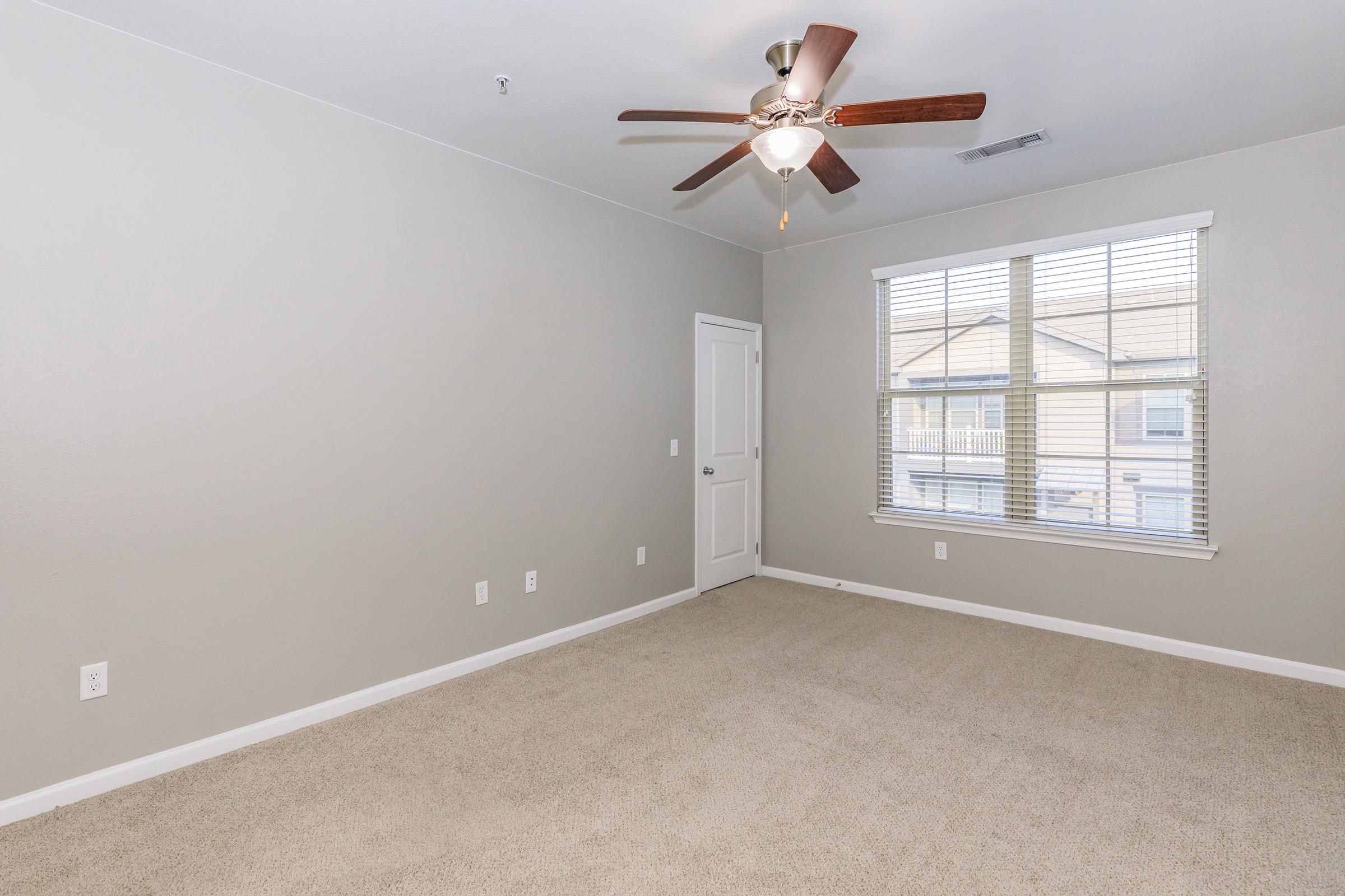 Spacious empty room with light beige carpet and light gray walls. A ceiling fan is mounted on the ceiling, and a large window with blinds allows natural light to fill the space. A door is visible on the side, leading to another area, enhancing the room's openness.