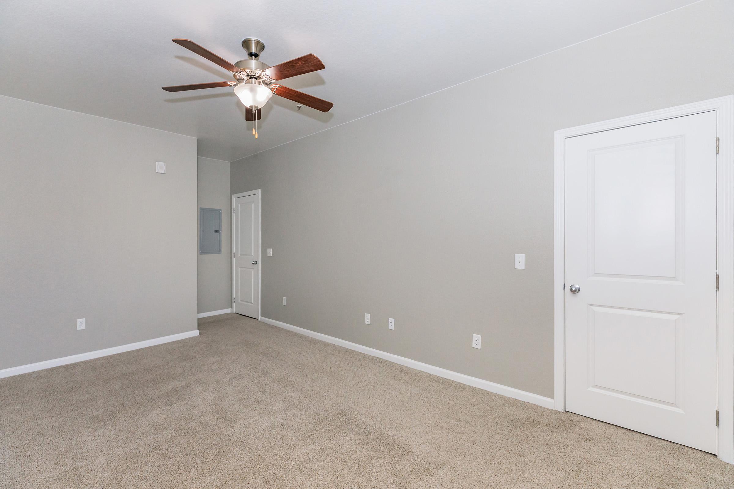 Empty room with light beige carpet, gray walls, and a ceiling fan. A white door on the right leads to another area, and a gray electrical panel is mounted on the wall. The space is bright and minimalistic, suitable for various uses or furniture arrangements.