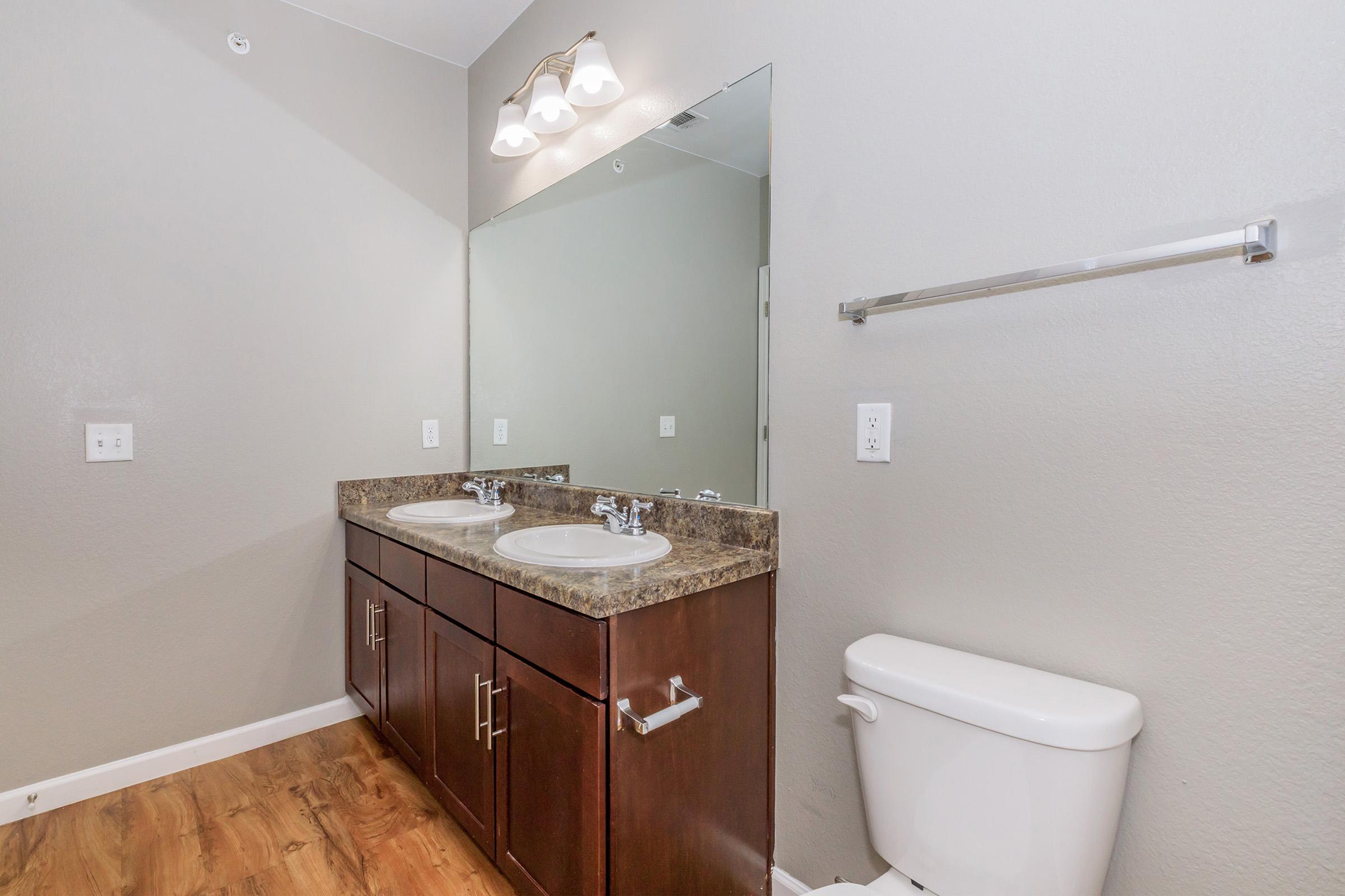 A modern bathroom featuring a double sink vanity with dark wood cabinetry, a large wall mirror, and a white toilet. The room has neutral-colored walls and wooden flooring, with bright lighting provided by a fixture above the sinks.