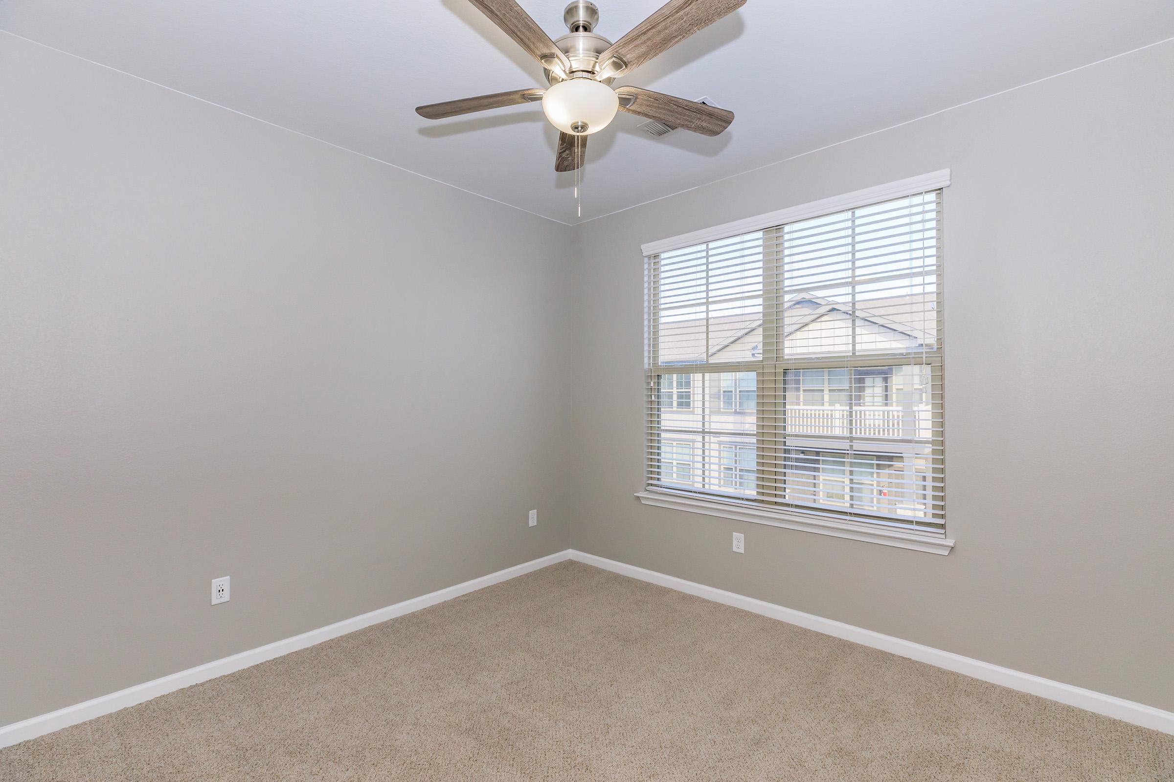 A vacant room featuring light gray walls, a ceiling fan with wooden blades, and a large window with blinds. The floor is covered in beige carpet, and there is a view of an exterior building outside the window.