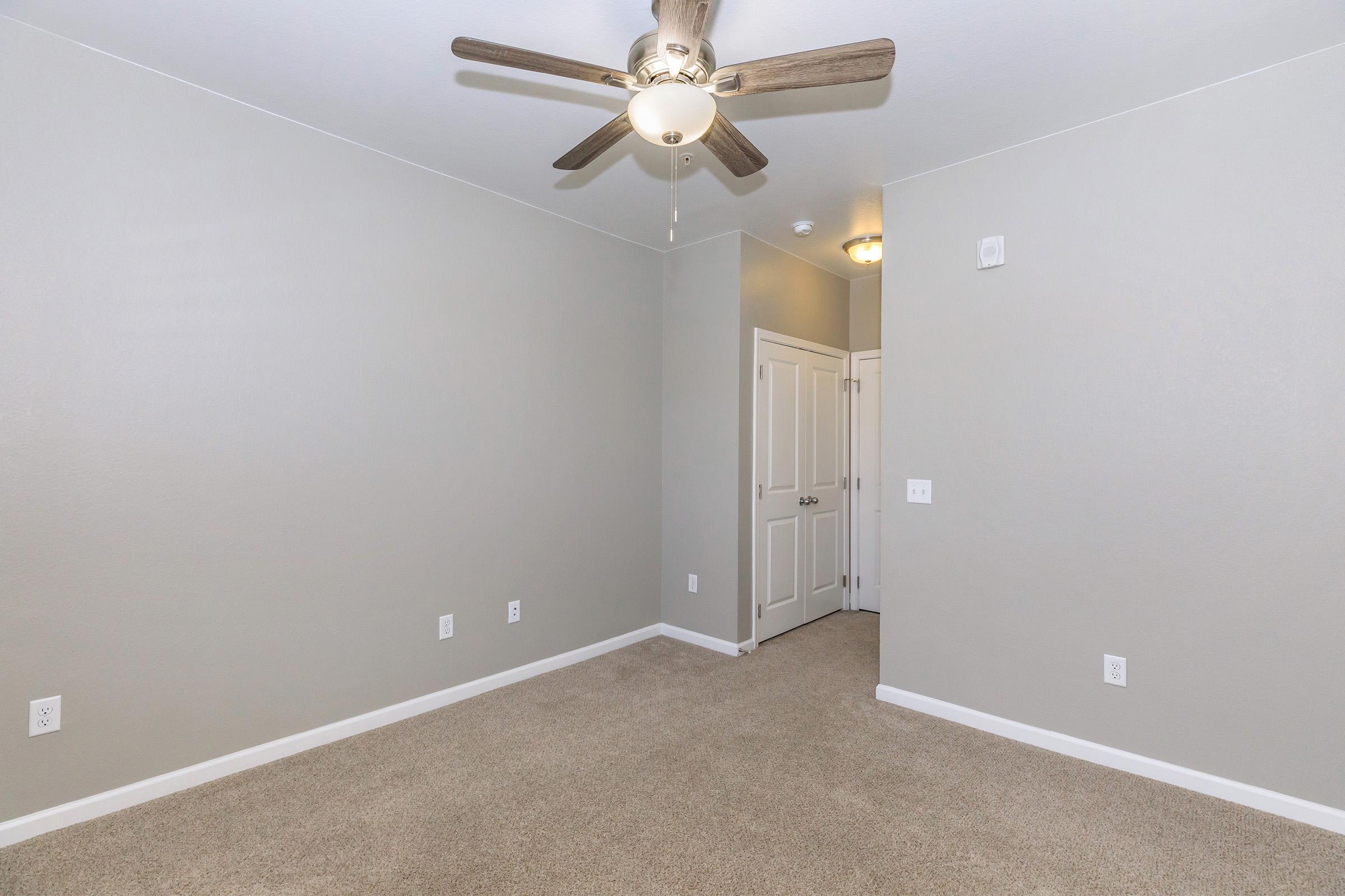 Empty room with light gray walls and a ceiling fan. The floor is covered in beige carpet. There are two doors on the right side, one leading to a closet and the other likely to a hallway. The ceiling has a light fixture illuminating the space. The room appears clean and freshly painted.