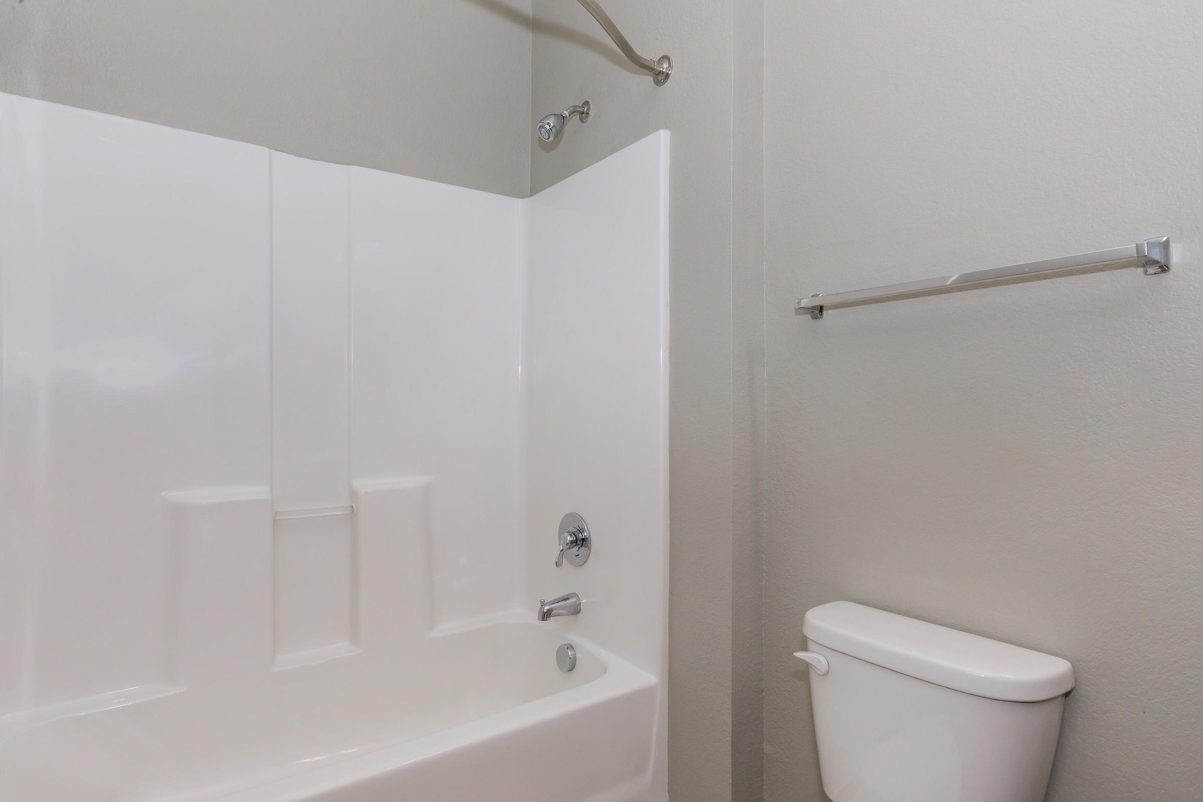 A clean and modern bathroom featuring a white shower stall with a chrome showerhead, a white toilet, and a towel bar on the wall. The walls are painted in a light gray color, creating a simple and spacious look.