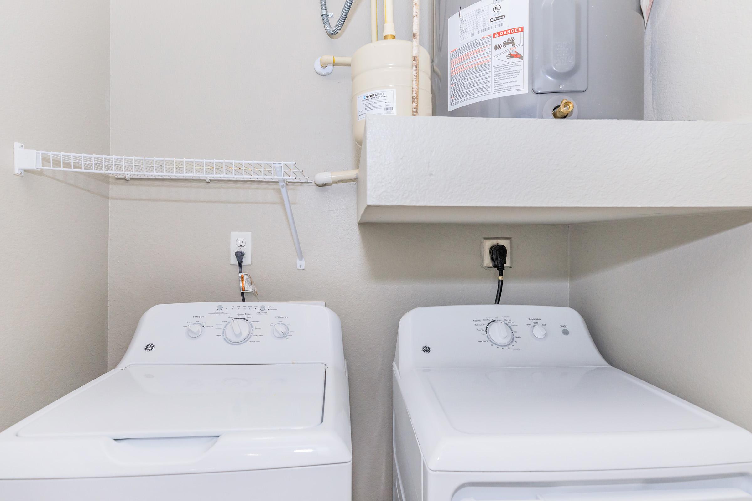 A laundry room featuring a washing machine and a dryer side by side. Above, a shelf holds a water heater. The walls are painted a light color, and there are electrical outlets and plumbing connections visible. The appliances are white and modern in style.