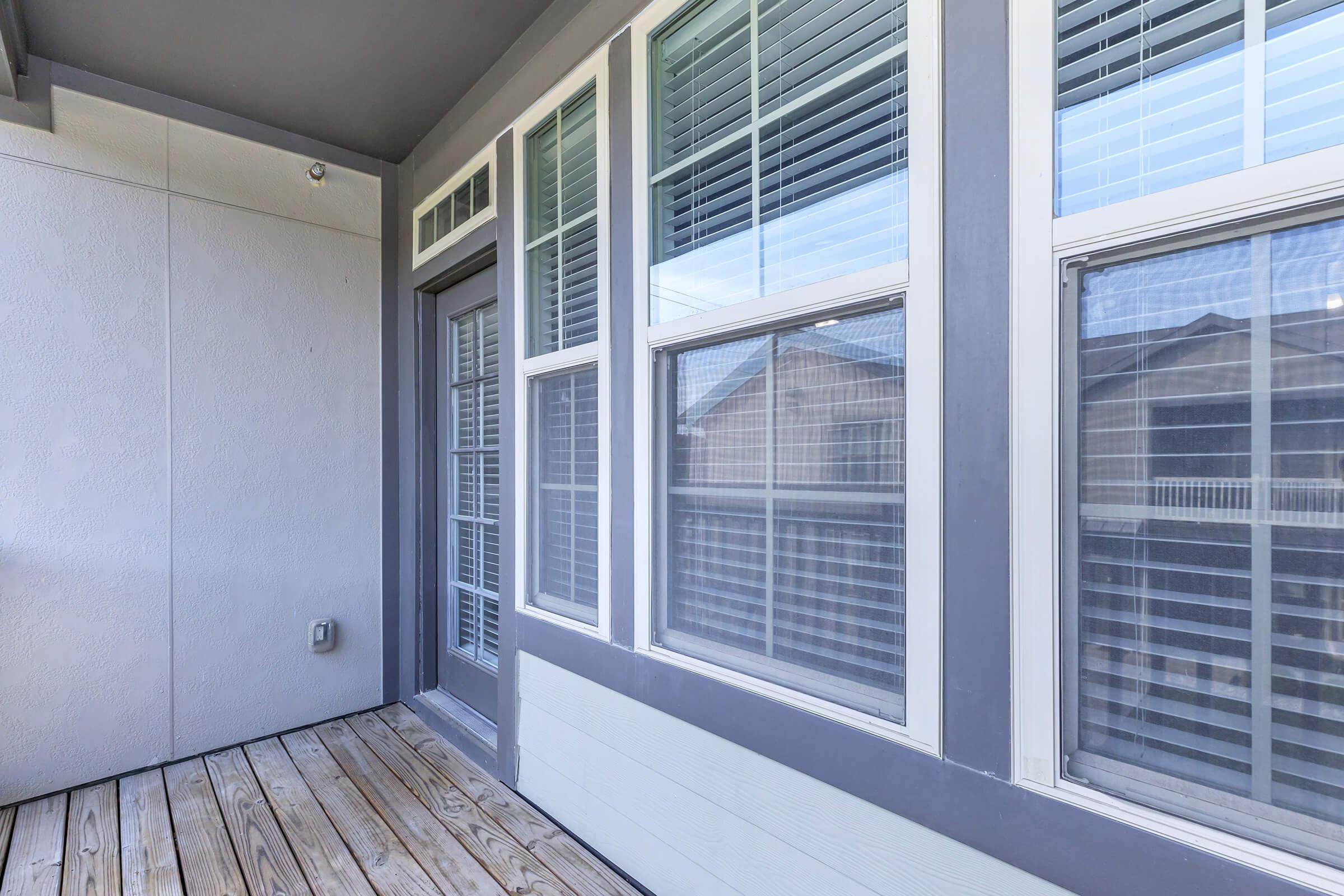 A corner of a balcony featuring wooden flooring and large windows with blinds. The walls are painted in a light color, and there's a small wall-mounted outlet visible. The overall design suggests a modern and cozy living space with plenty of natural light.