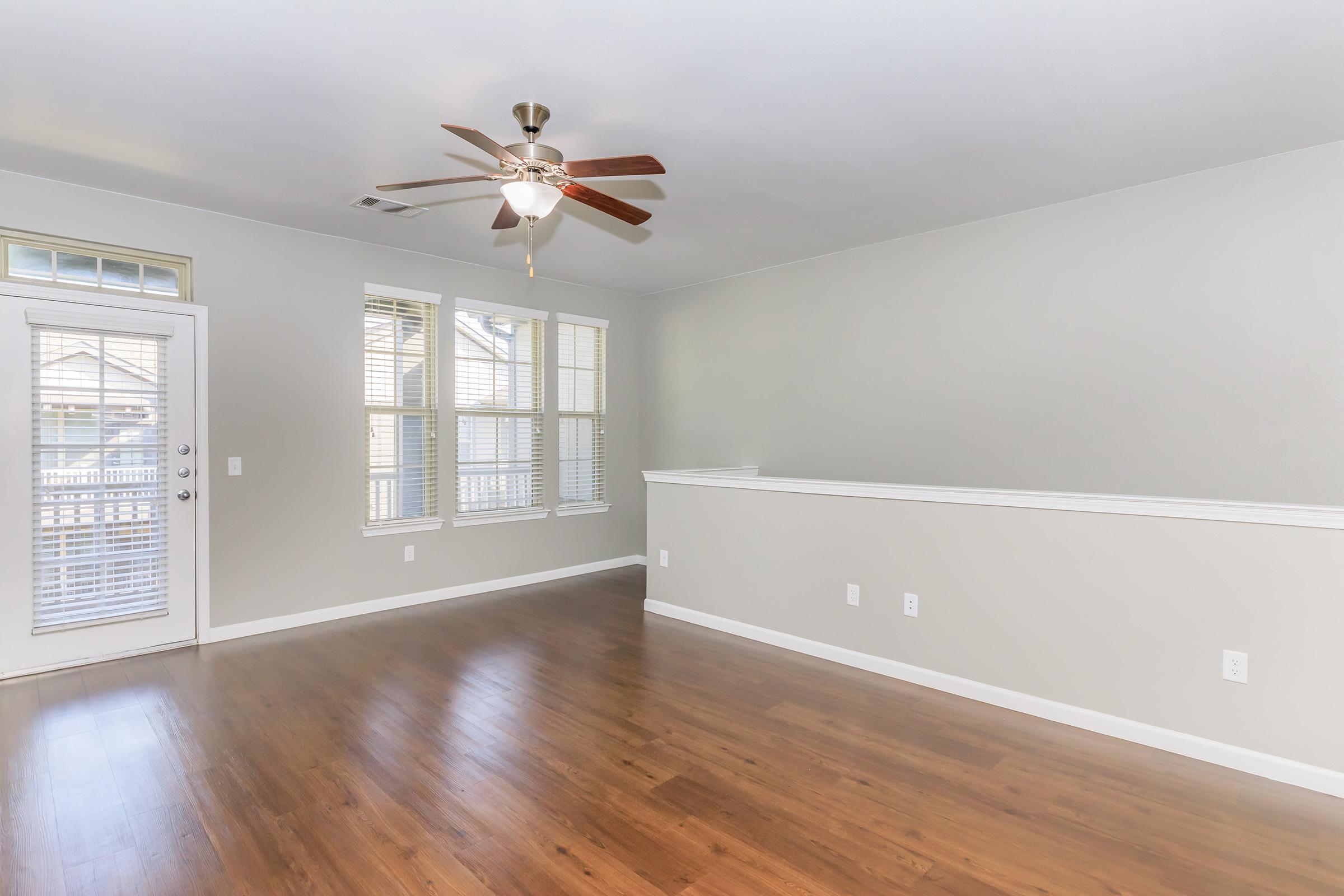 Spacious, well-lit living room featuring light brown wooden flooring, a ceiling fan, and large windows with blinds. The walls are painted a soft gray, and there is a door leading outside. A decorative half wall adds dimension to the space.