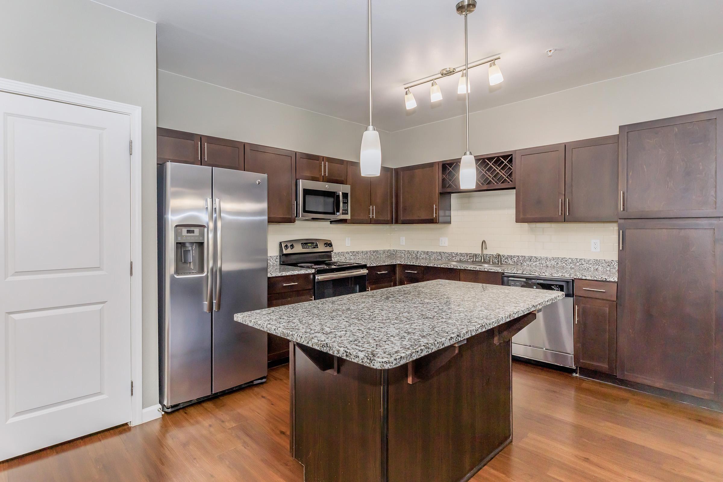 Modern kitchen featuring dark wood cabinetry, stainless steel appliances, a large central island with a granite countertop, and pendant lighting. The space is well-lit and includes a microwave, oven, and dishwasher, with a clean and contemporary design.