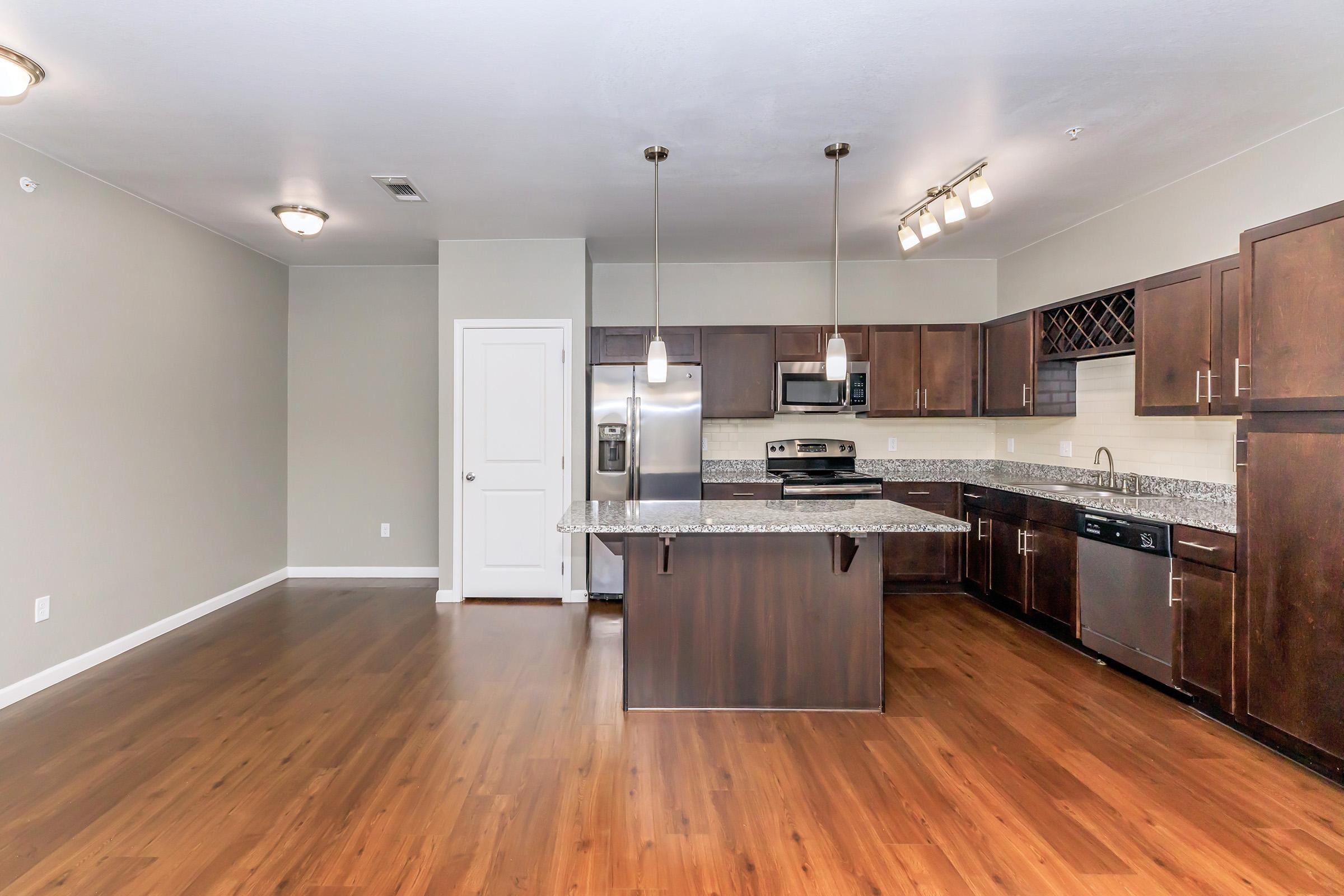 Modern kitchen interior featuring dark wooden cabinets, granite countertops, stainless steel appliances, and an island with seating. The space has hardwood flooring and neutral-colored walls, with pendant lighting above the island and a door leading to another room.