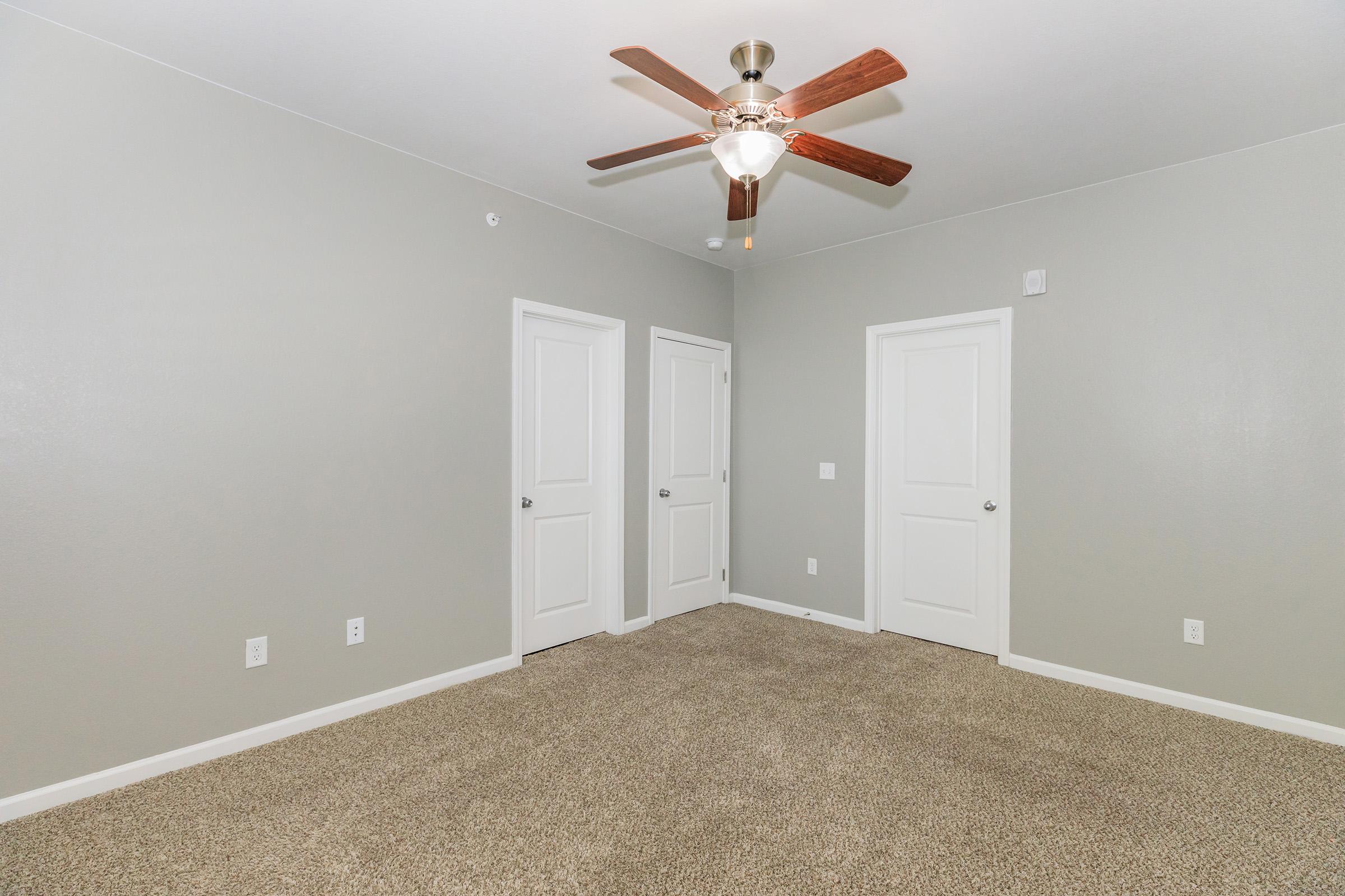 A spacious, light gray room featuring a ceiling fan with wooden blades. There are two closed doors on the left and one on the right, with a soft beige carpet covering the floor. The walls are bare, creating a clean and simple atmosphere.