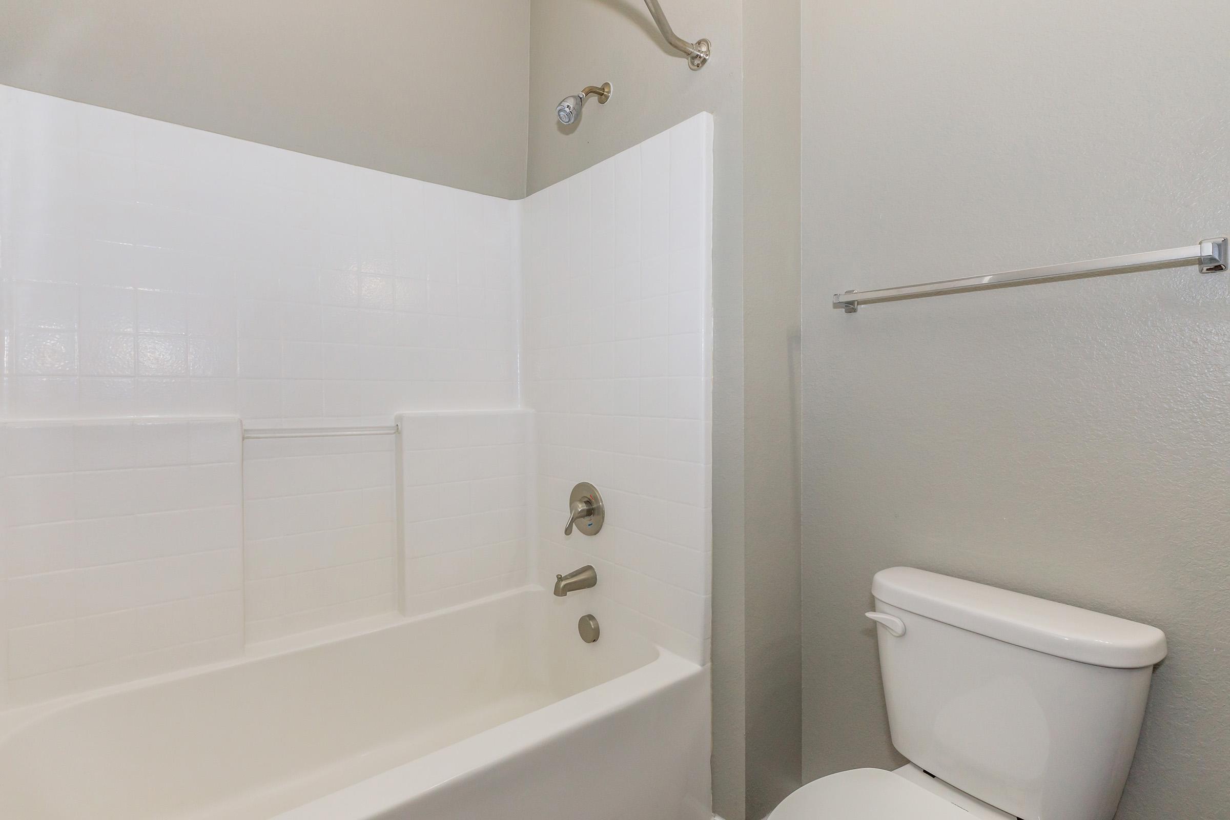 A clean and simple bathroom featuring a white bathtub with a showerhead, white tiled walls, a chrome faucet, and a chrome towel bar. A white toilet is positioned next to the bathtub. The walls are painted in a light color, contributing to a bright and open atmosphere.