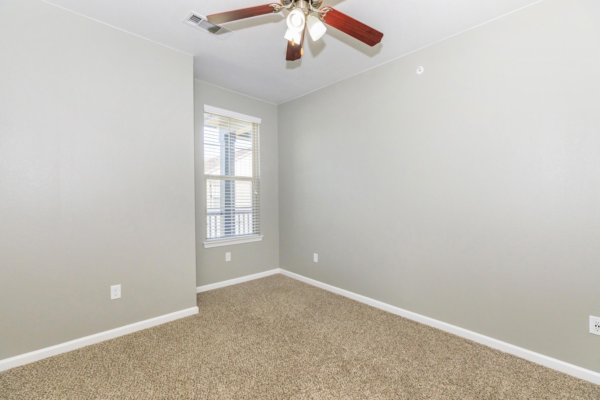Empty room with light gray walls and carpeted floor. A ceiling fan with wooden blades is installed. There are two windows on the left wall, allowing natural light to enter, and minimal decor, creating a spacious and clean atmosphere.