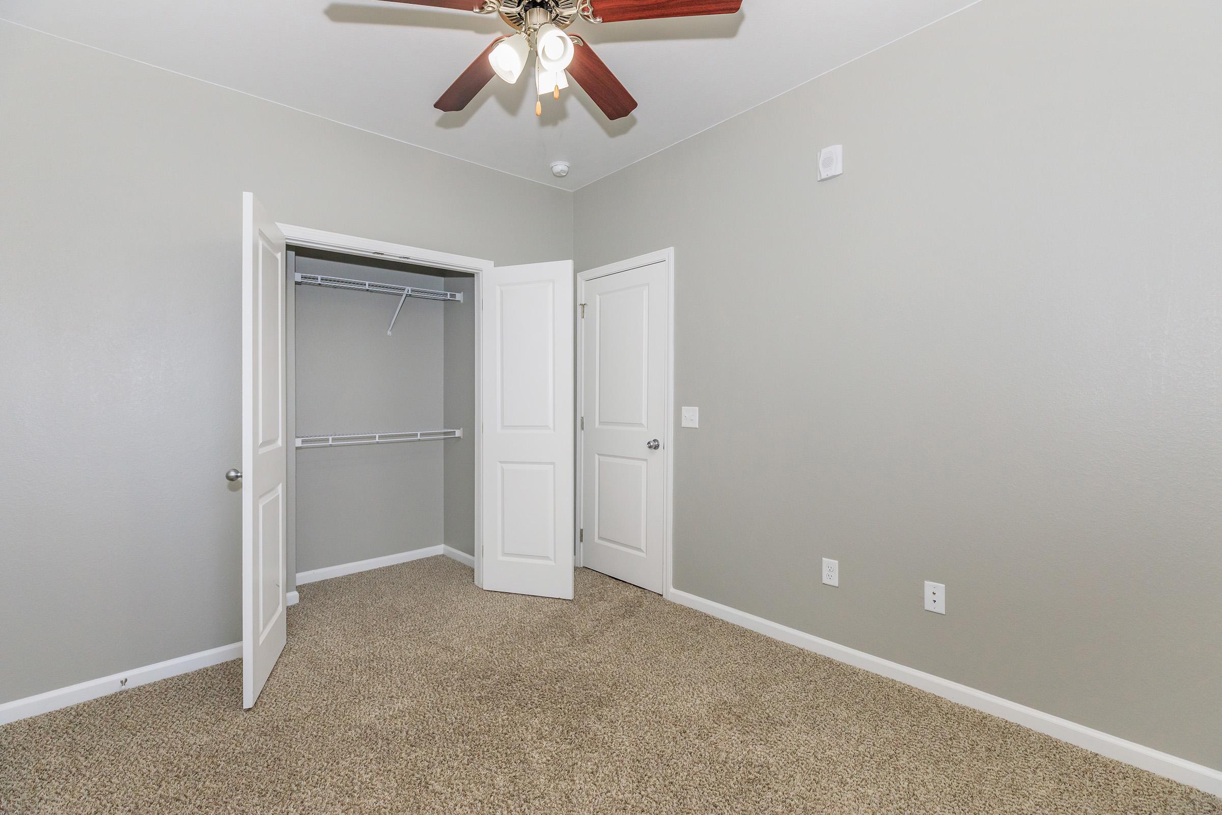 A well-lit, empty bedroom featuring light gray walls, a ceiling fan with four lights, and plush carpet. The room has two doors, one leading to a closet with shelving, and another door leading outside. The overall space is clean and ready for furniture placement.