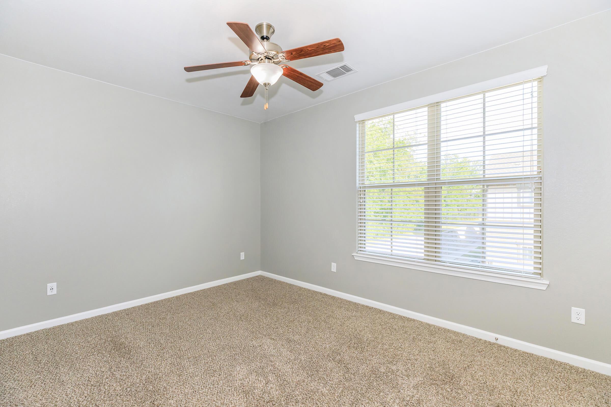 Empty room with light gray walls and a ceiling fan with wooden blades. The floor is covered in beige carpeting, and there's a large window with blinds allowing natural light to enter. The room is spacious and uncluttered, making it suitable for various purposes.