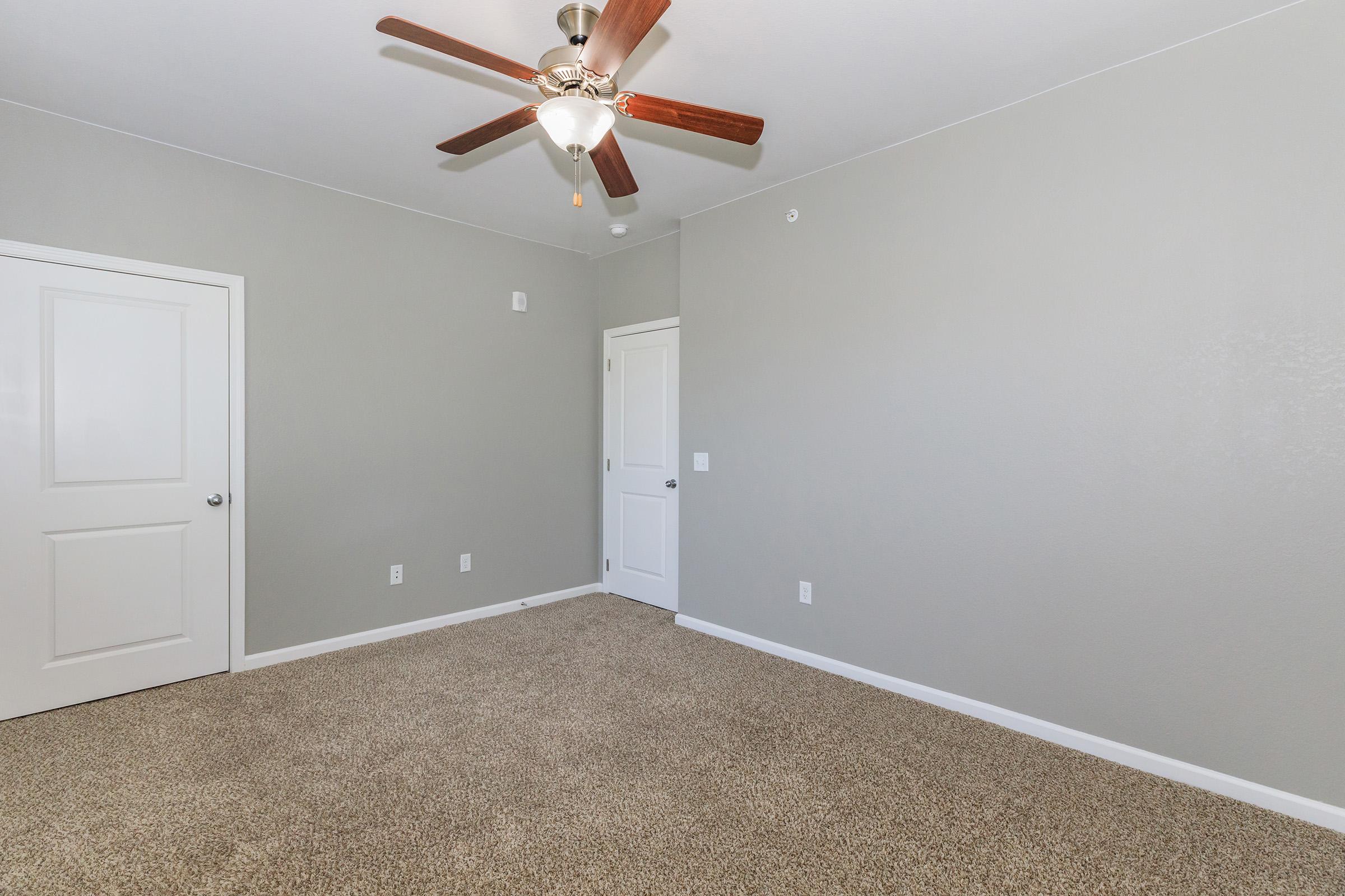 Empty room with light gray walls and carpeted flooring. A ceiling fan with wooden blades is mounted on the ceiling. There are two white doors on opposite walls, one leading to a closet. The room has a bright, uncluttered appearance, suitable for various uses.