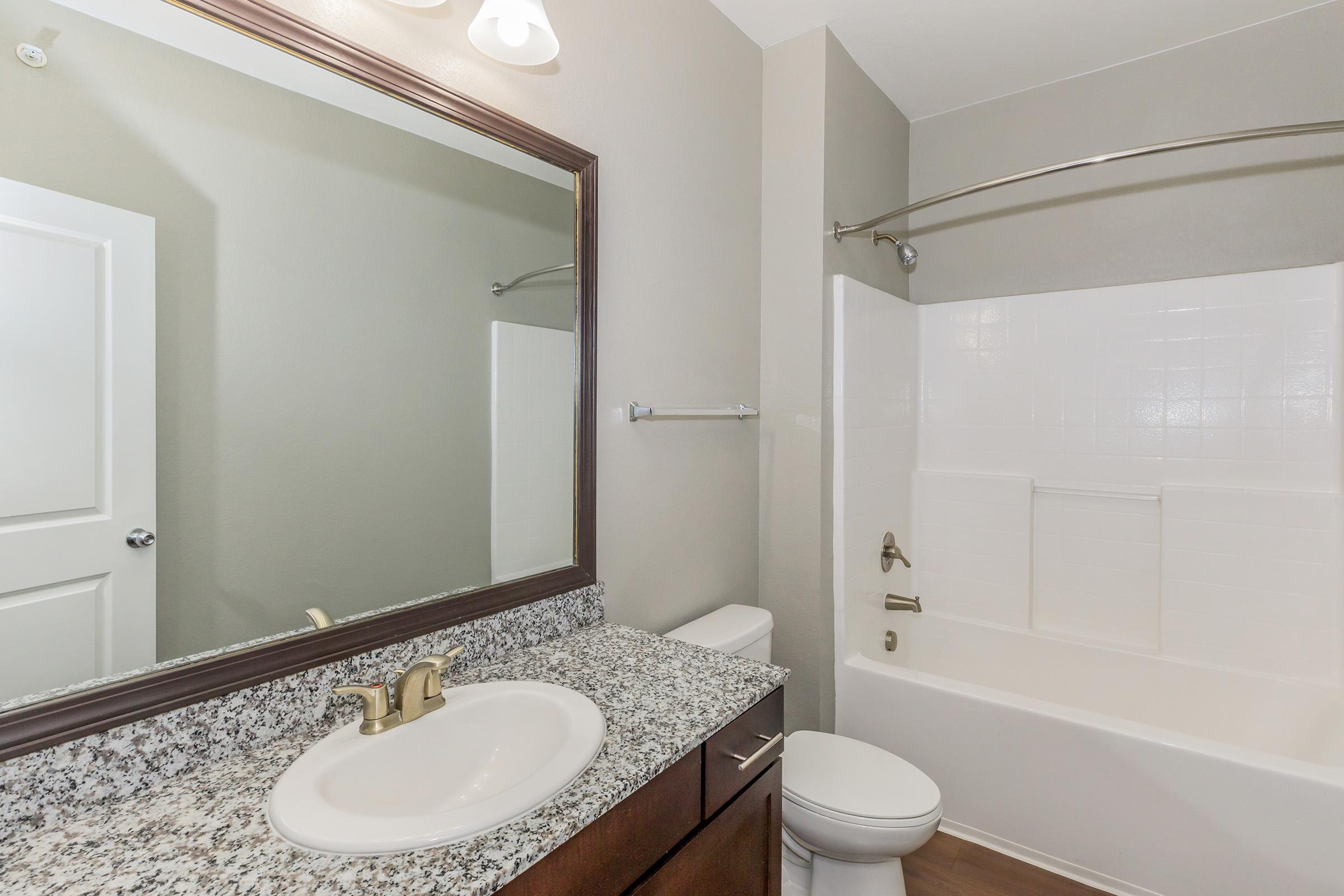 A clean and modern bathroom featuring a white bathtub with a shower, a granite countertop vanity with a sink, a large mirror, and neutral-colored walls. The space has a contemporary feel, with wooden flooring and standard bathroom fixtures.