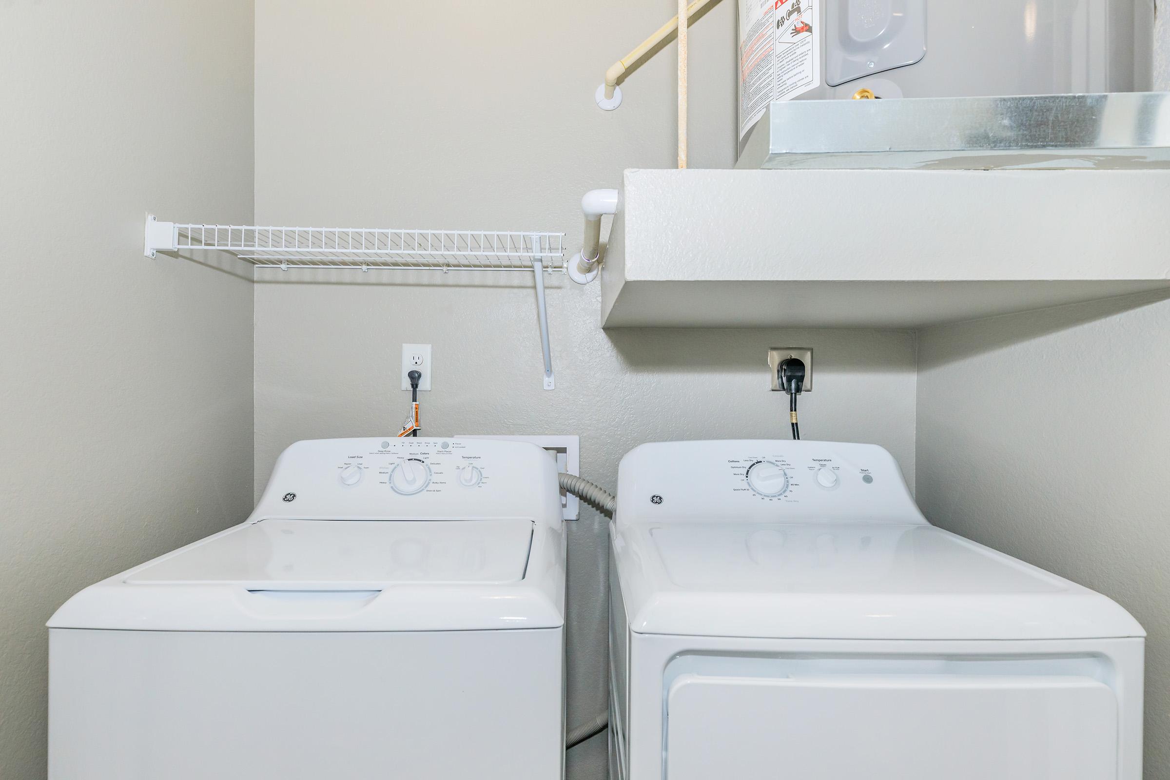 A laundry room featuring a white washing machine and dryer side by side, with a wall-mounted shelf above. The walls are painted a light color, and there's a water heater visible in the corner. Electrical outlets are located near the appliances, providing power for their operation.