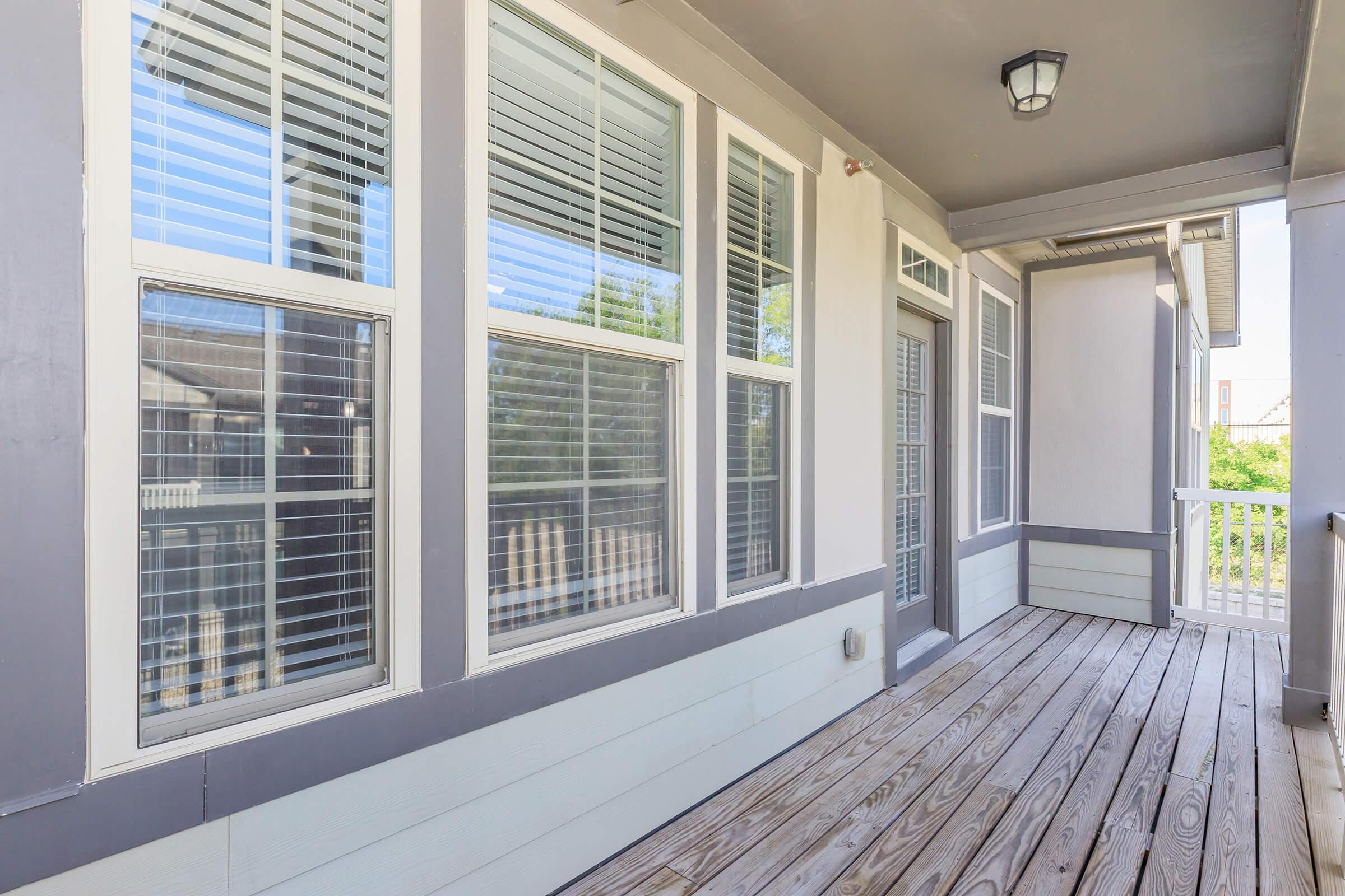 A spacious balcony with wooden flooring, featuring multiple large windows with white blinds. The exterior walls are painted in light grey and white, creating a modern look. A light fixture hangs from the ceiling above. Greenery is visible outside through the windows, adding a touch of nature to the scene.