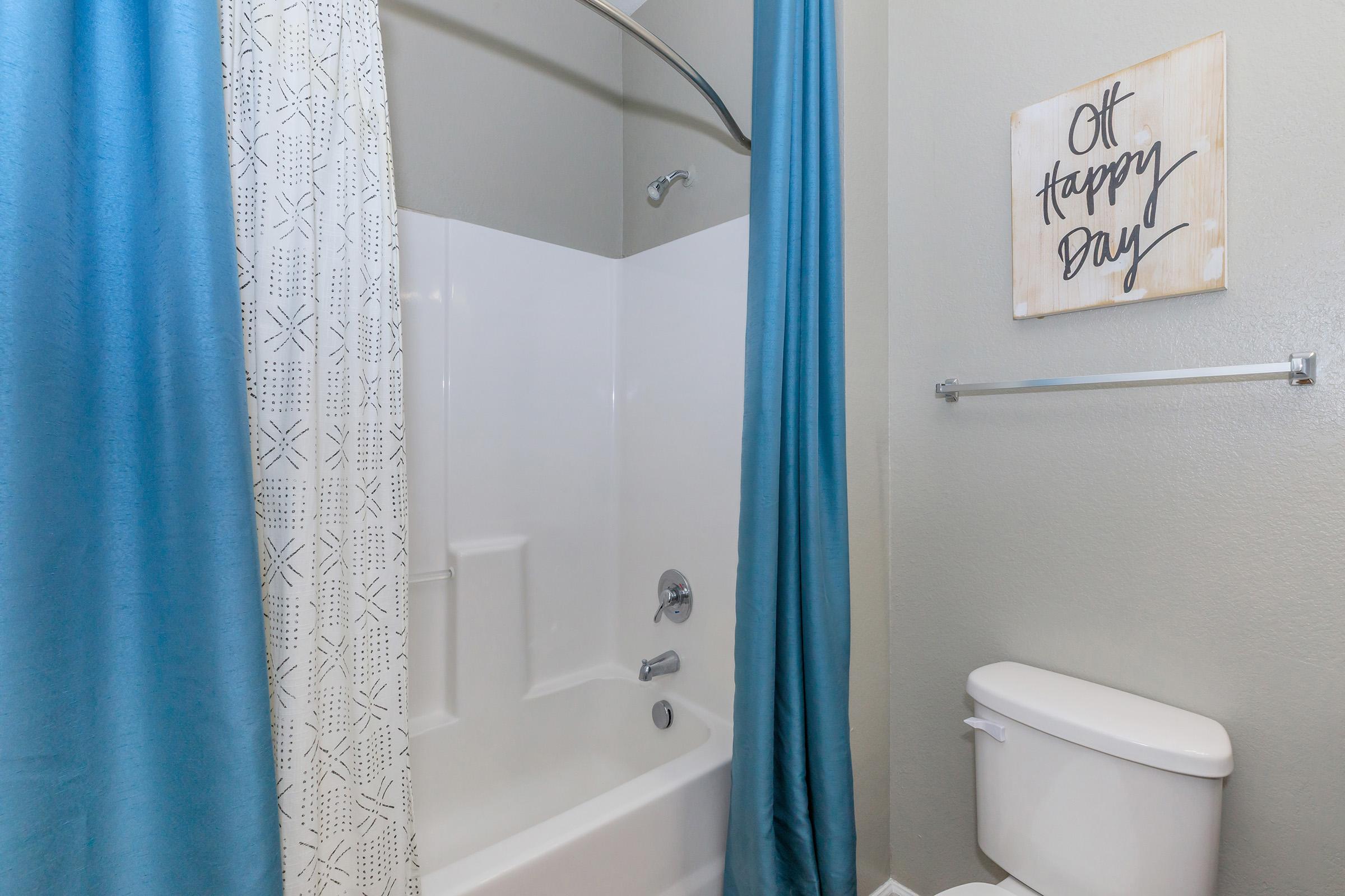 A bathroom featuring a bathtub with a built-in shower, a blue shower curtain, and a white toilet. On the wall, there is a framed sign that reads "Oh Happy Day." The walls are painted in a light gray color, creating a calm atmosphere.