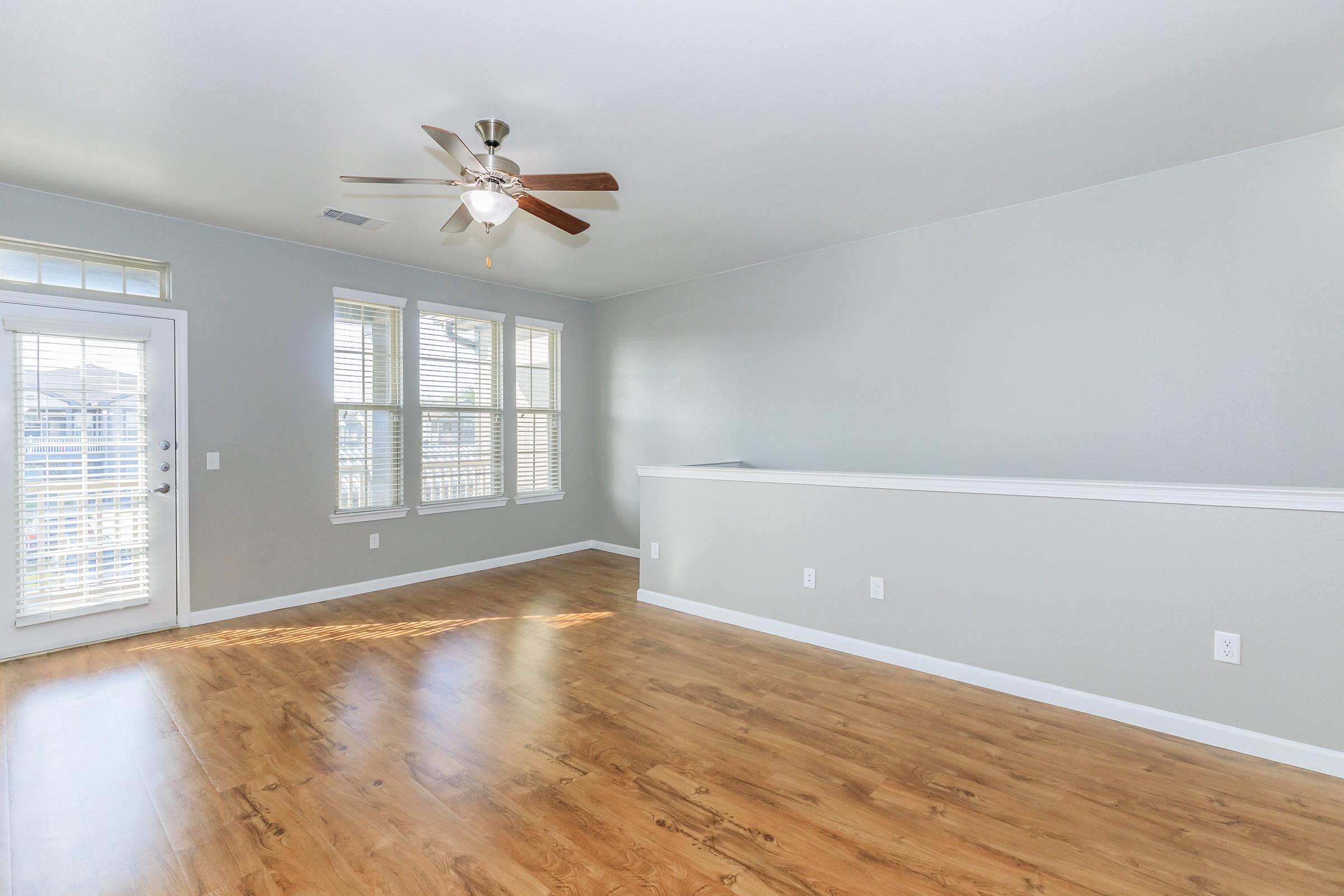 Bright, spacious living room with light gray walls and wooden laminate flooring. Features a ceiling fan, large windows with blinds allowing natural light, and a sliding glass door leading to a balcony or patio. The room is empty, creating a fresh and inviting atmosphere.