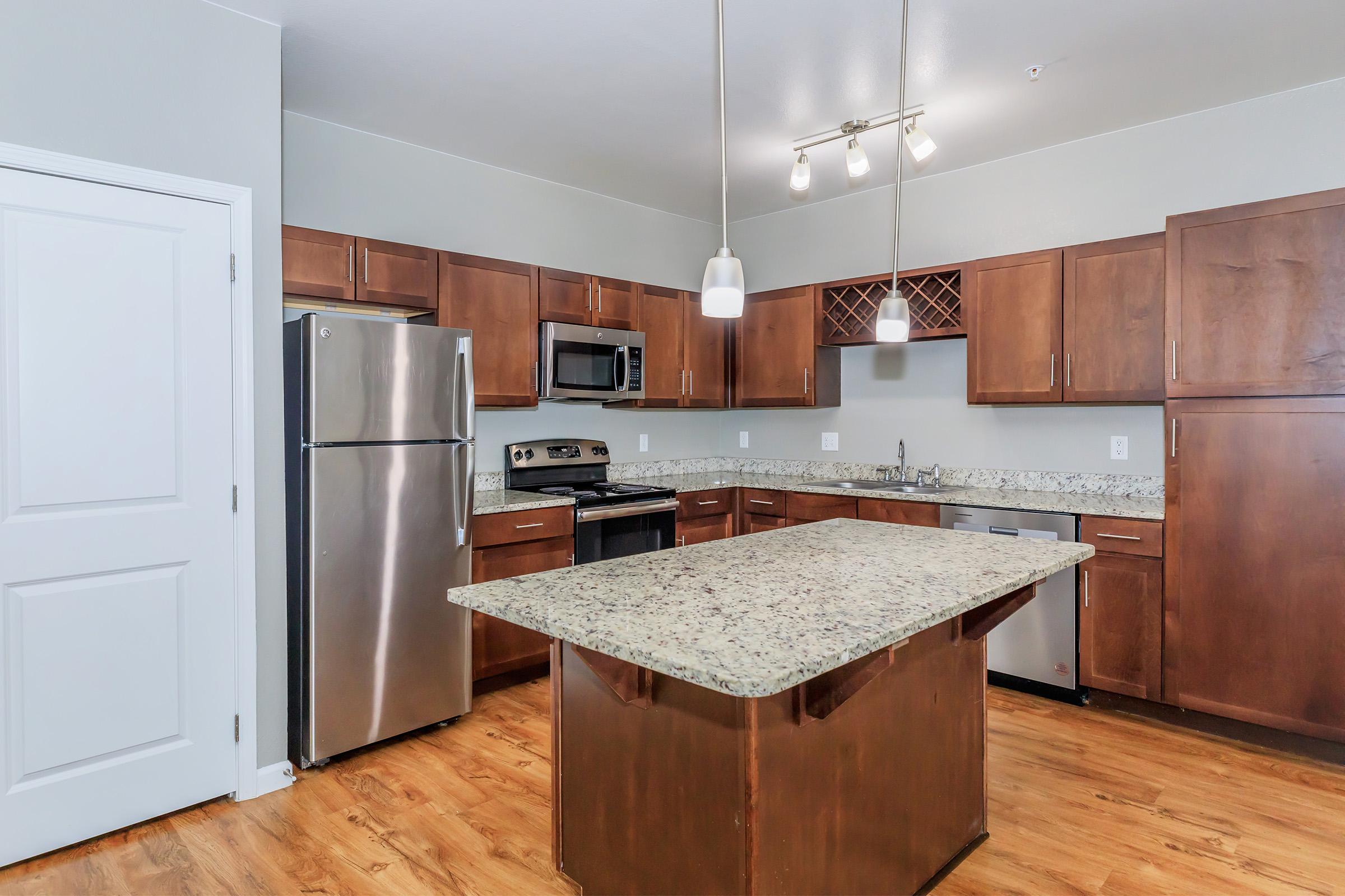 Modern kitchen featuring wooden cabinetry, granite countertops, stainless steel appliances including a refrigerator, oven, and microwave, along with a central island. The space is well-lit with pendant lighting and has a wine rack above the sink area. The floor is hardwood, contributing to a warm atmosphere.