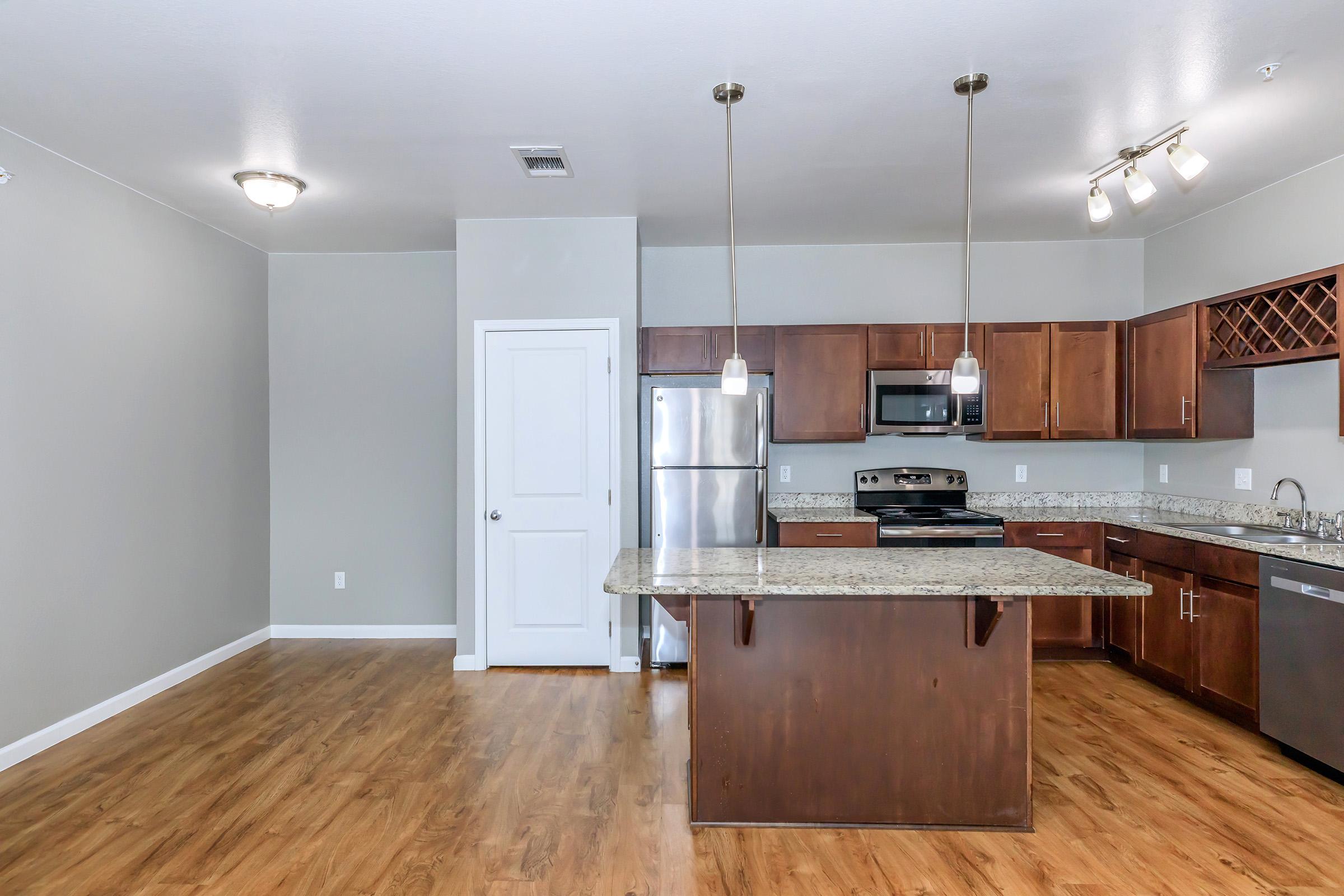 Modern kitchen featuring wooden cabinetry, stainless steel appliances, and a large island with a granite countertop. The space includes a door leading to another area, warm wood flooring, and neutral-colored walls. Soft lighting fixtures above the island and a wine rack integrated into the cabinets add a stylish touch.