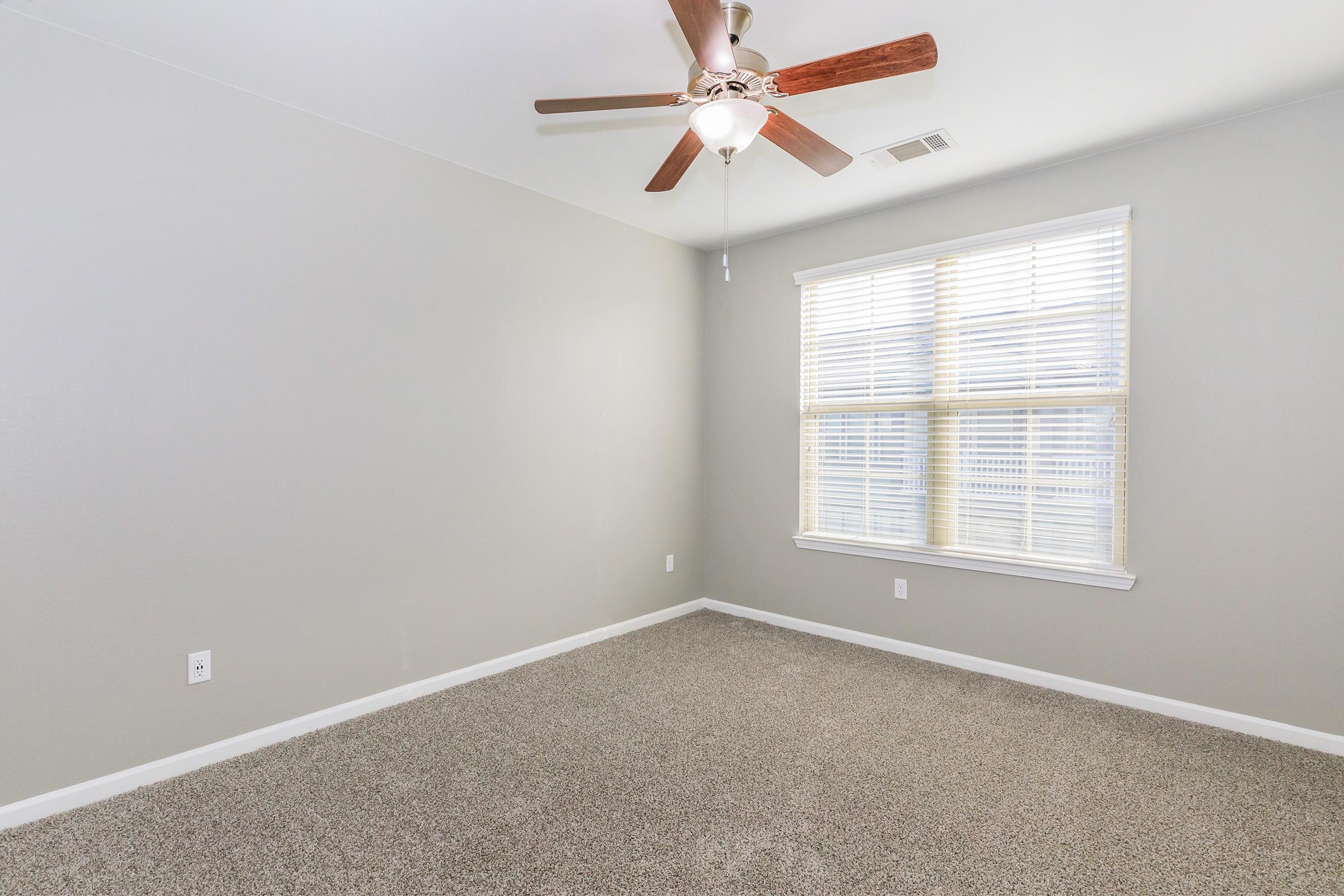 Empty room with light gray walls, a ceiling fan with wooden blades, and a large window featuring blinds. The floor is covered in beige carpeting, creating a cozy atmosphere. Natural light filters through the window, illuminating the space.
