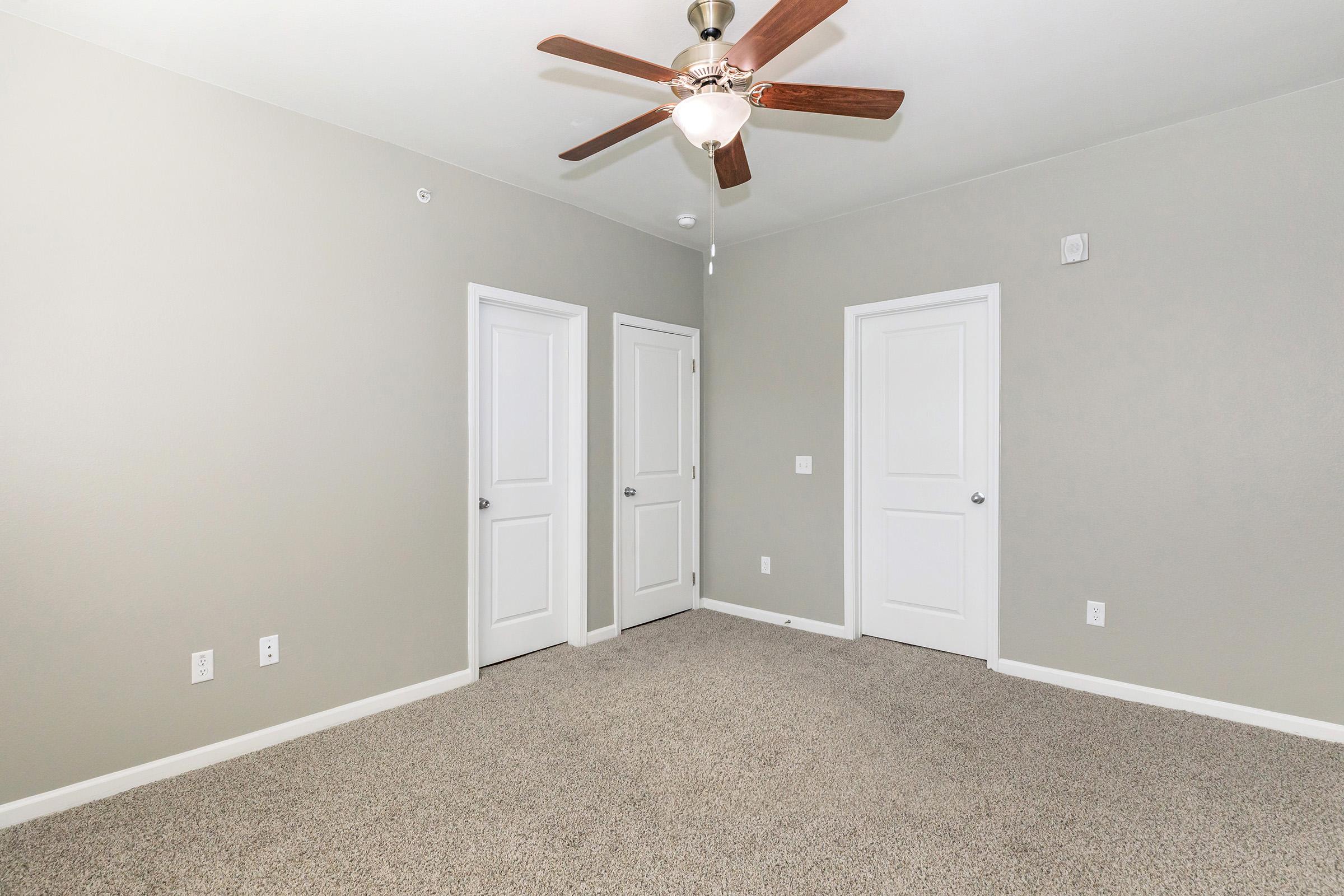 A neutral-colored room with light gray walls and carpet flooring. It features a ceiling fan with wooden blades and three closed white doors along the walls. The space is uncluttered, creating a simple and modern aesthetic.