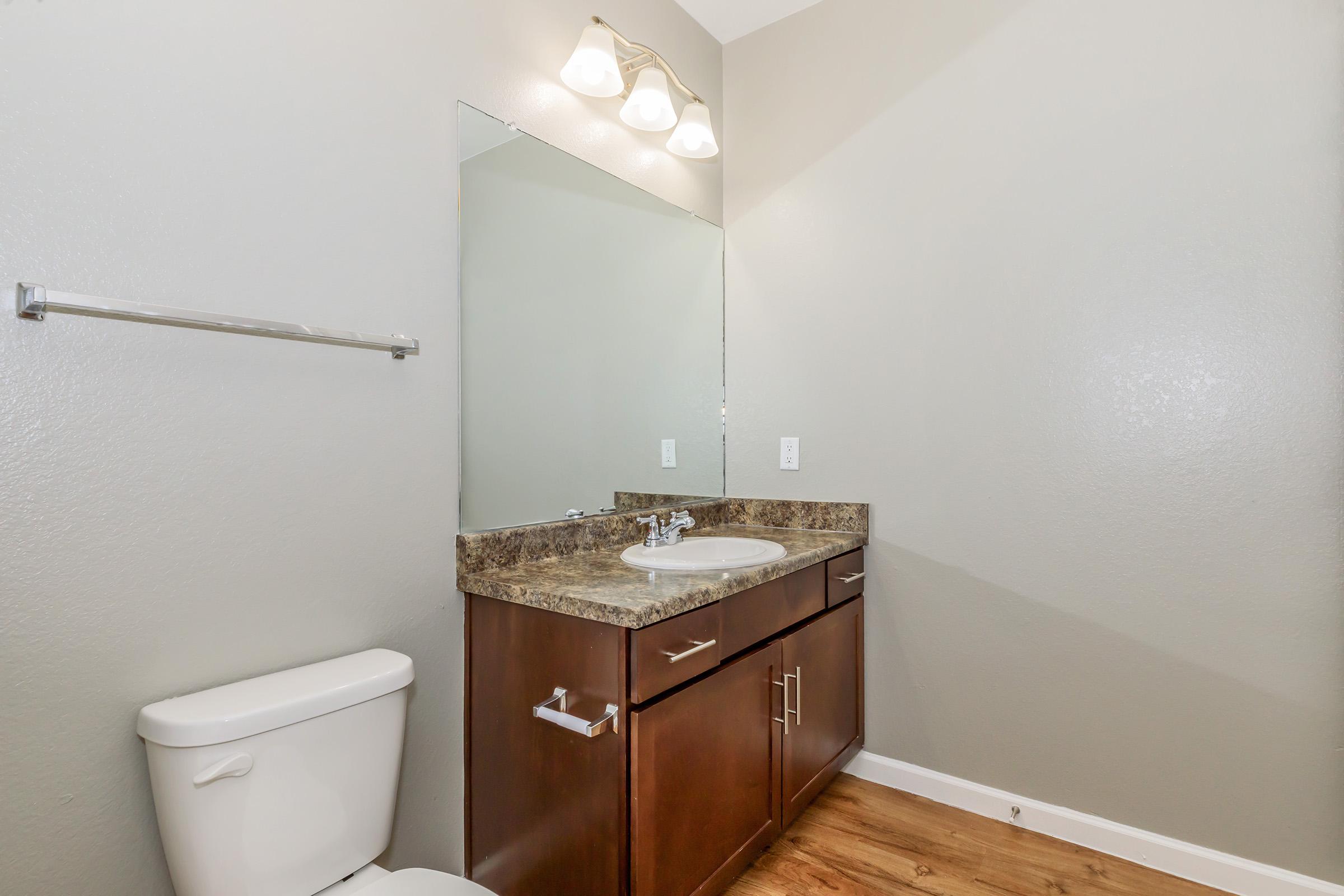 A modern bathroom featuring a toilet, a vanity with a granite countertop, a sink, and a large mirror. Soft lighting is provided by three wall-mounted fixtures. The walls are painted in a light gray color, and the floor has a wood-like appearance.