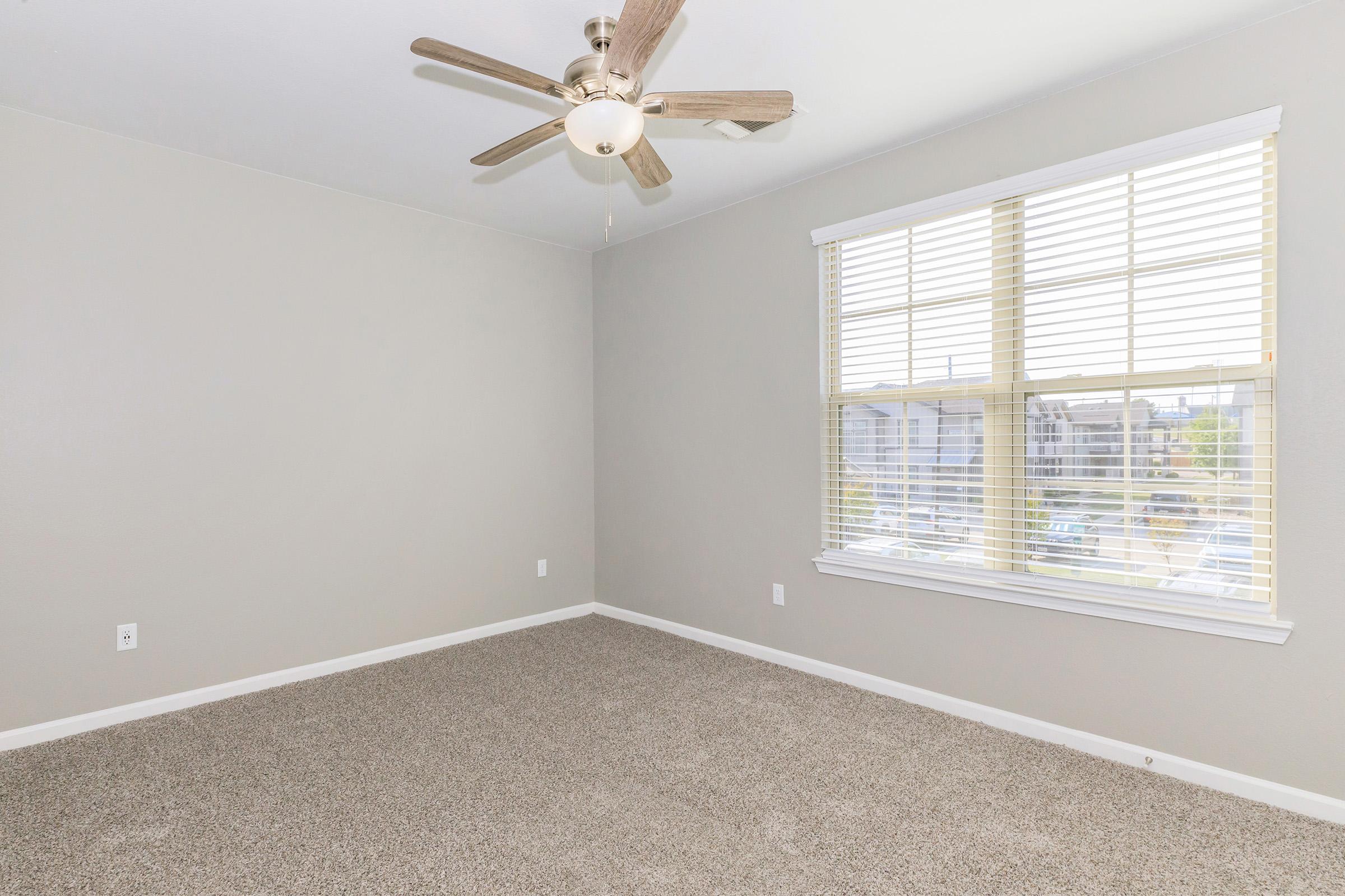 A spacious, empty room with light gray walls and a ceiling fan. The floor is covered in tan carpet. A large window with white blinds lets in natural light, revealing a view of a parking area outside. The room is unfurnished, creating a bright, open atmosphere.