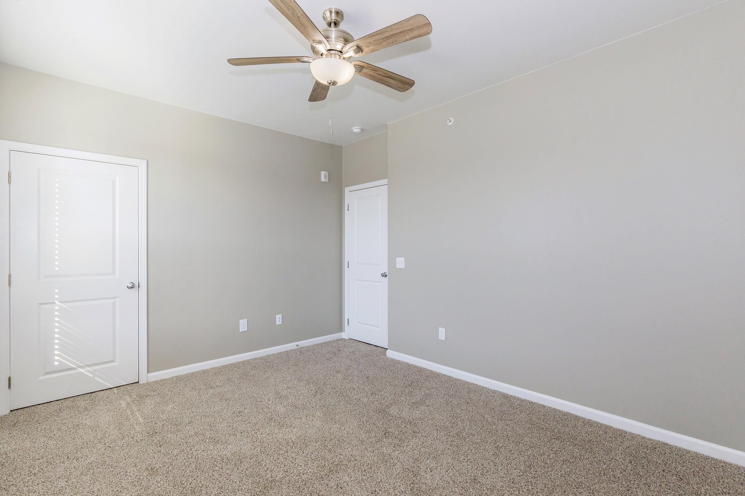 A room with light gray walls and carpeted flooring. It features a ceiling fan with wooden blades, a closed white door, and a partially open door leading to another space. The room is empty, creating a spacious and clean appearance. Natural light may enter through the unseen window.