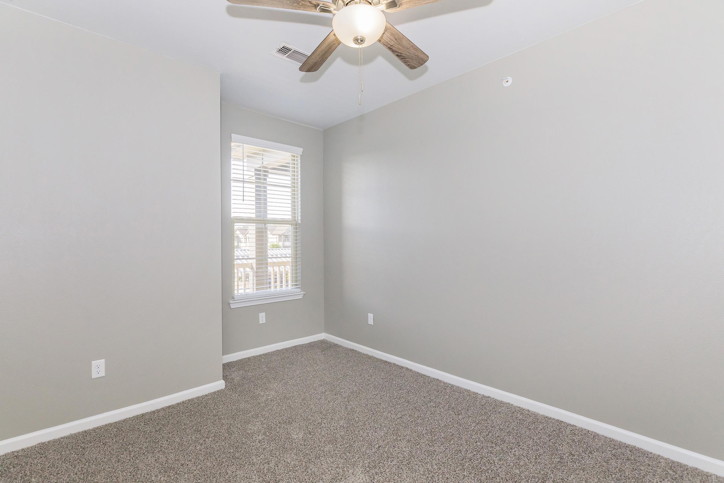 Empty room with light gray walls, a ceiling fan, and a window with white blinds. The floor is covered in beige carpet, and the space has natural light coming in from the window.