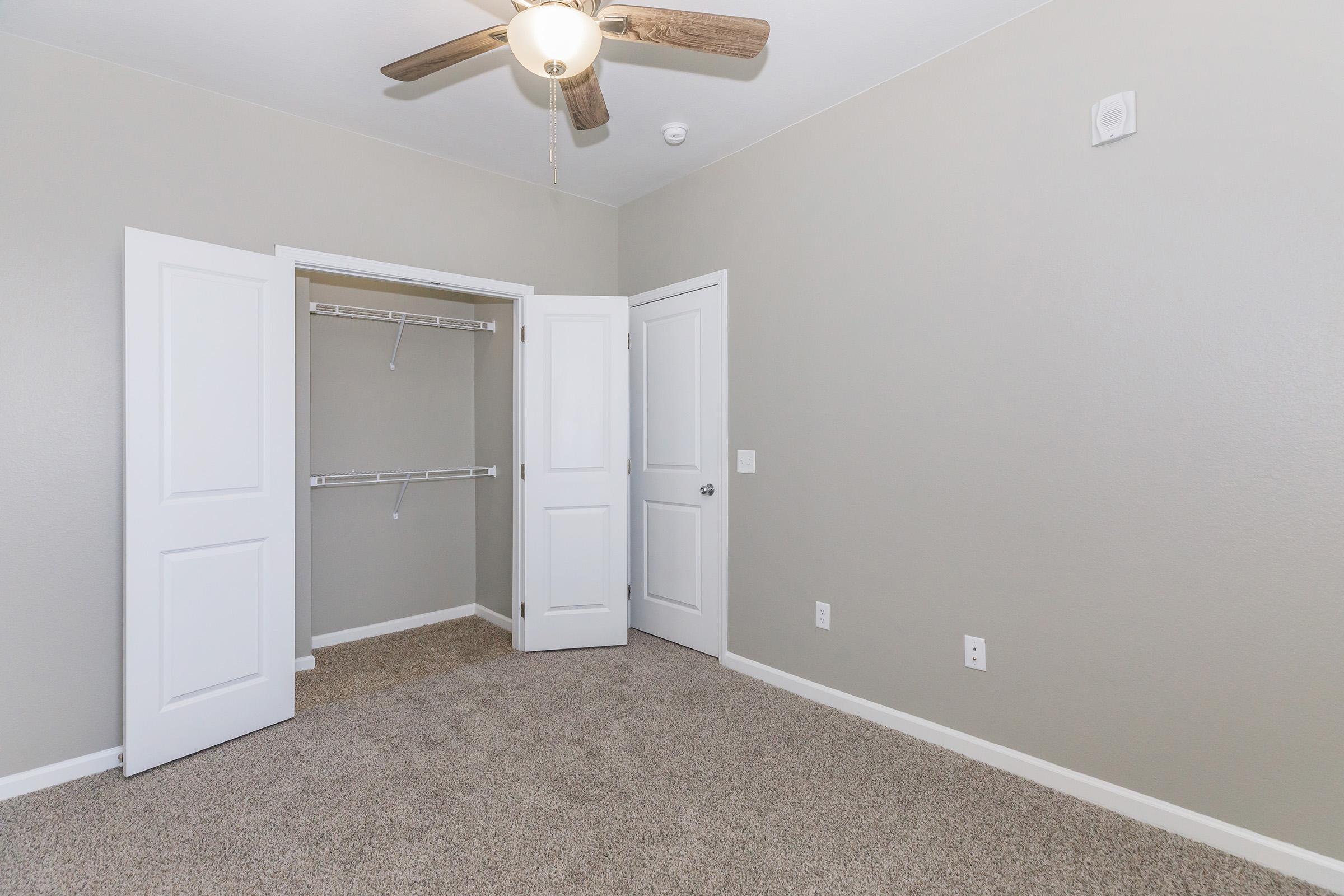 A vacant bedroom featuring light gray walls, a ceiling fan with wooden blades, and plush carpet. The room has two open white doors leading to a closet with shelving, and there is a small white wall-mounted feature. The overall atmosphere is bright and inviting.