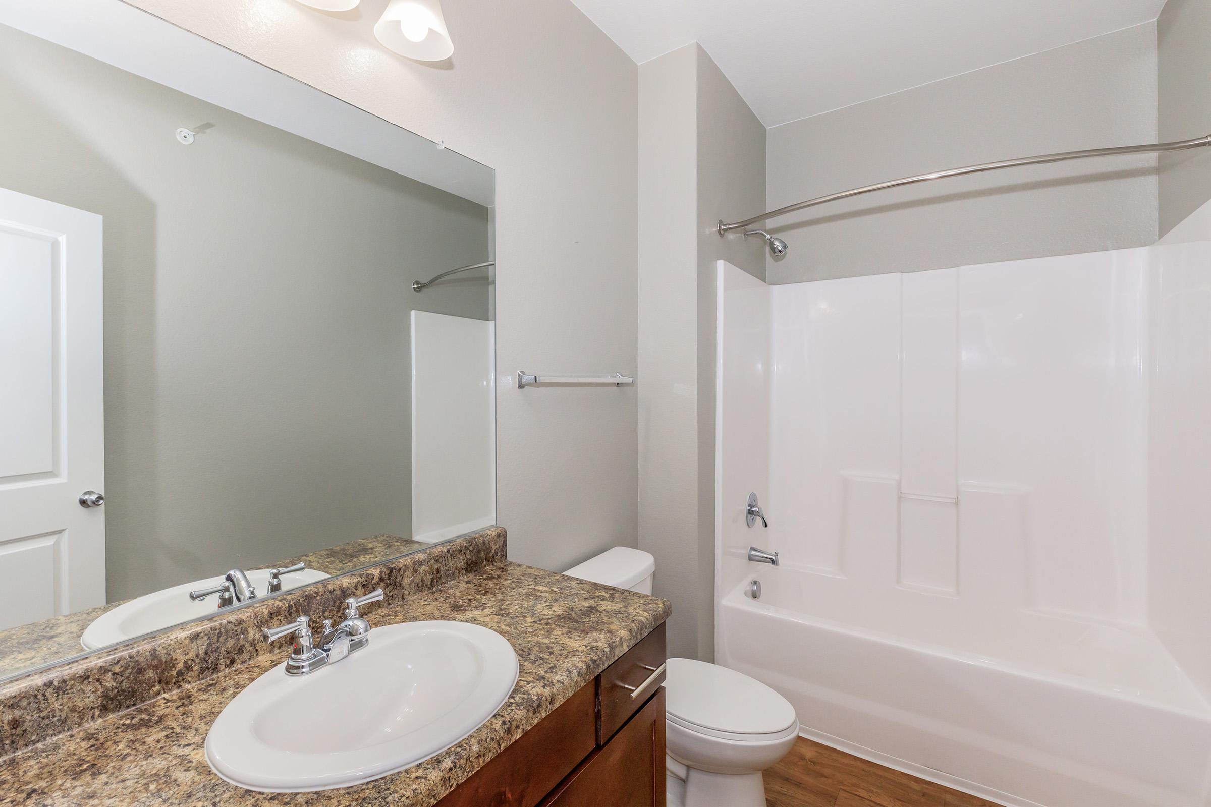 A modern bathroom featuring a large mirror above a granite countertop sink, a white bathtub with a shower curtain rod, and a toilet. The walls are painted in a light color, and the flooring appears to be wood laminate. Bright lighting fixtures are installed on the ceiling.
