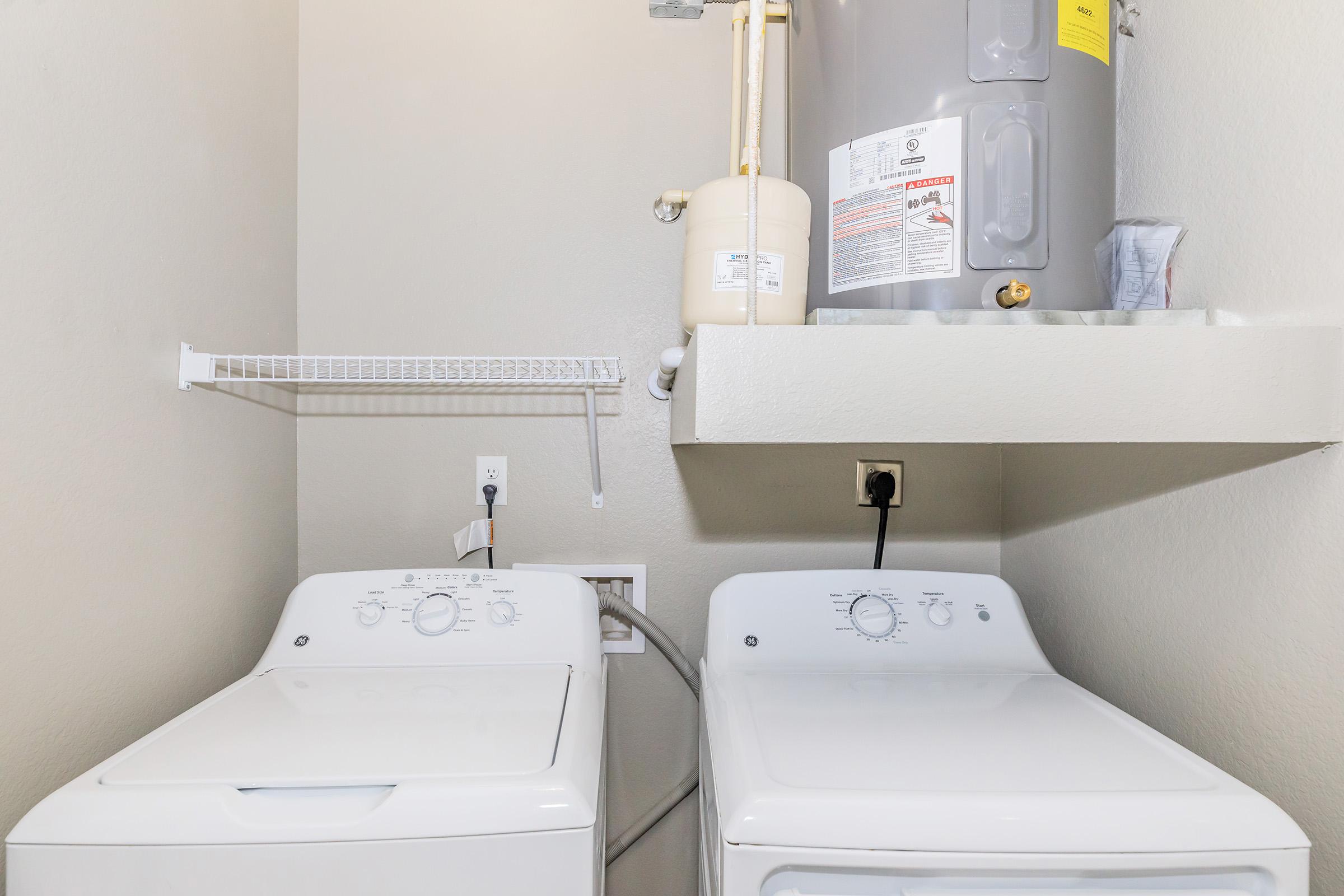 A laundry room featuring a top-loading washing machine and a dryer side by side. Above the appliances, there is a shelf for storage and a water heater mounted on the wall. The walls are painted in a light color, creating a clean and tidy appearance.