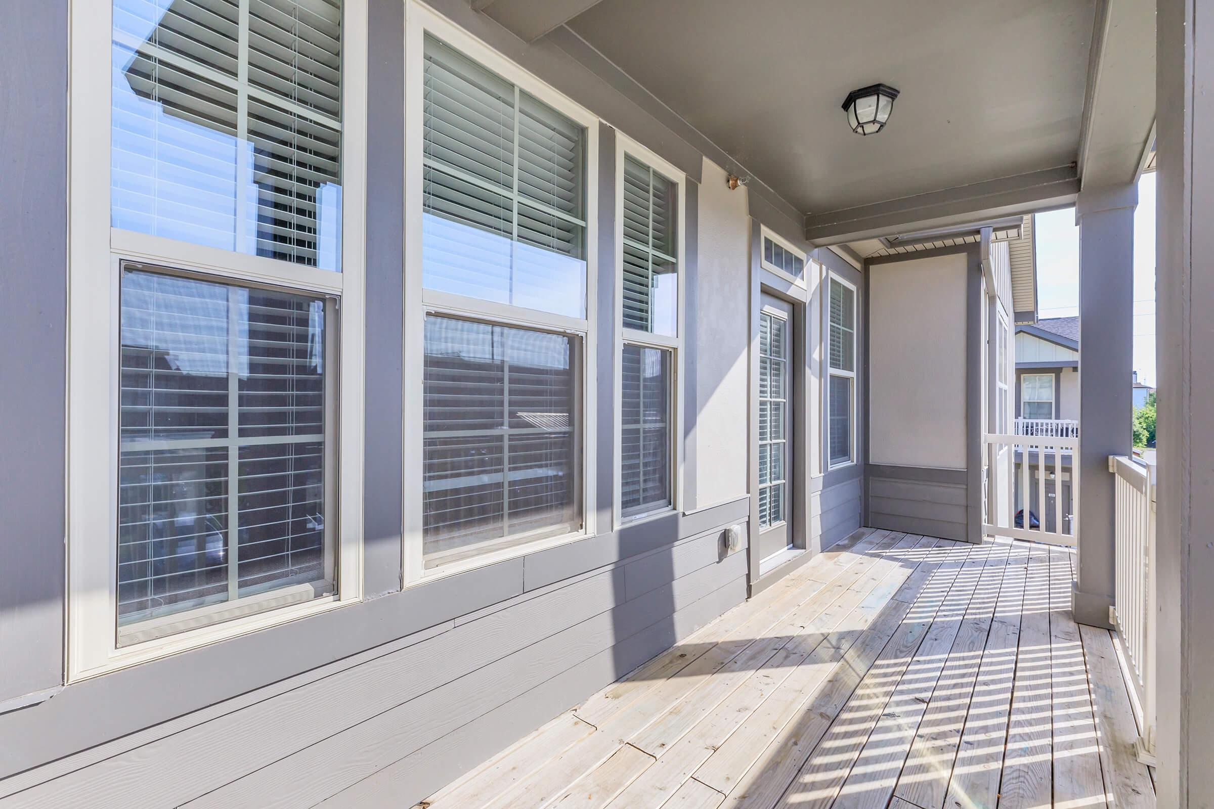 A well-lit balcony with wooden flooring and large windows featuring white blinds. The walls are painted in light grey, and there's a ceiling light fixture visible. The view from the balcony shows a neighboring building and clear skies.