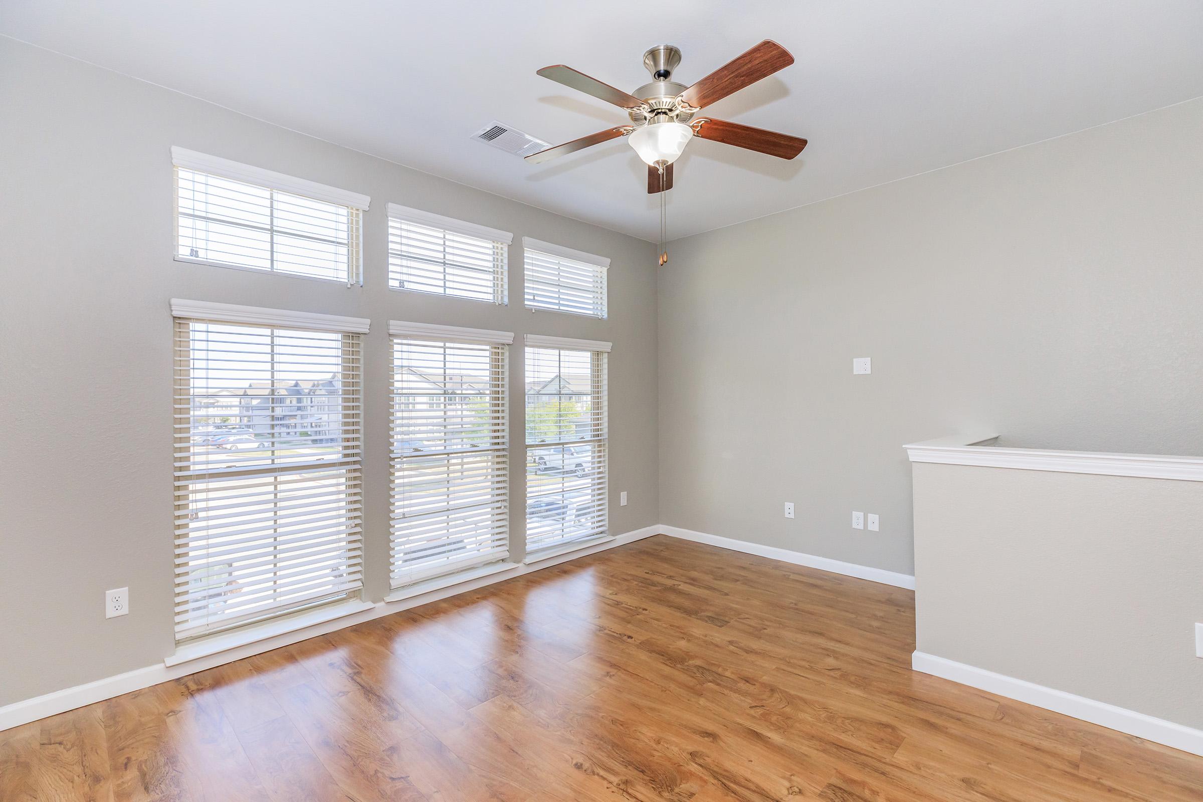A spacious, well-lit room featuring large windows with blinds, a ceiling fan, and light brown wooden flooring. The walls are painted in a neutral gray color, and there is a low wall in the background. The room has a clean and modern aesthetic, ideal for various uses.