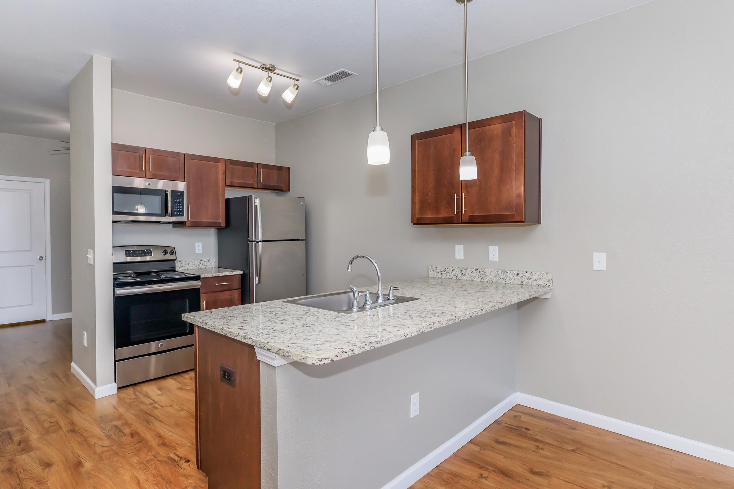 Modern kitchen featuring granite countertops, stainless steel appliances, and wooden cabinetry. Pendant lights hang above a spacious island with a sink. The open layout connects the kitchen to a living area with hardwood flooring and neutral wall colors.