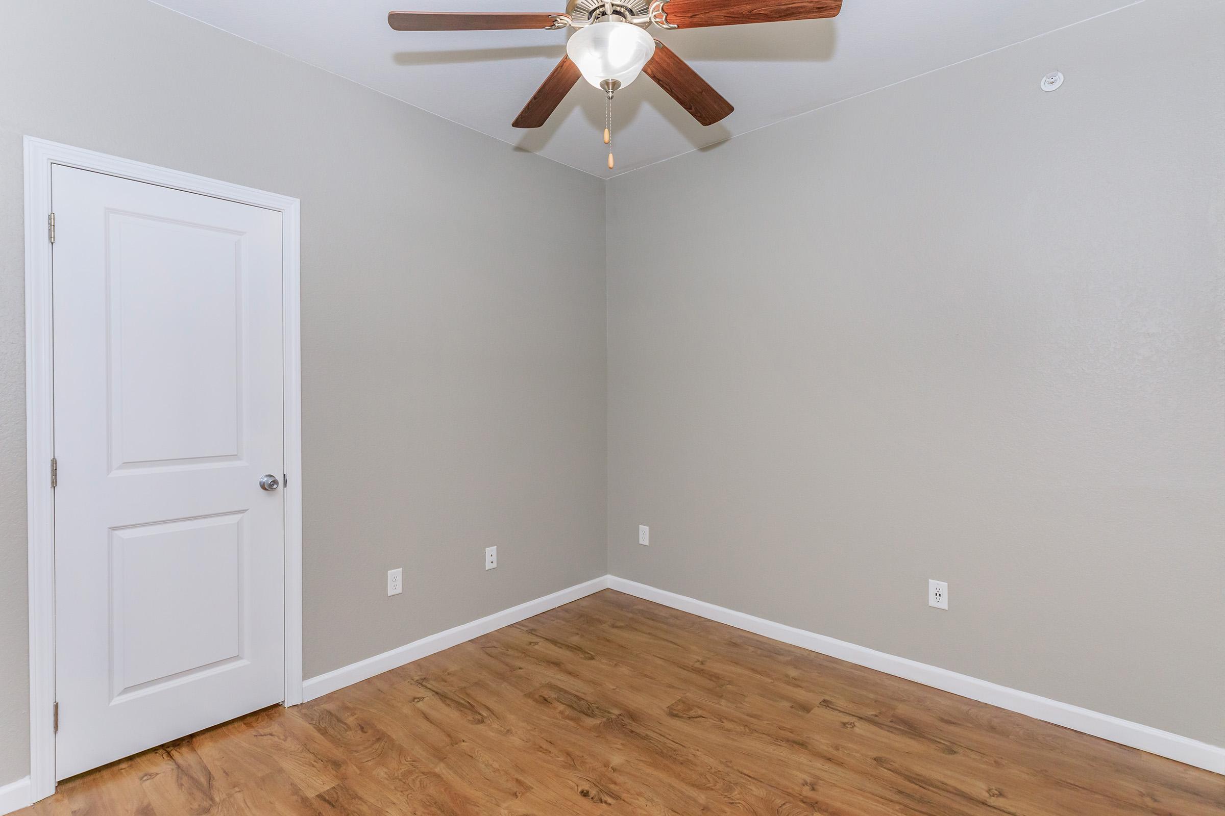 Empty room featuring light gray walls and a ceiling fan with wooden blades. The floor is a laminate wood-style, and there is a white door on the left side. The space is bright and uncluttered, ideal for various uses or decoration.