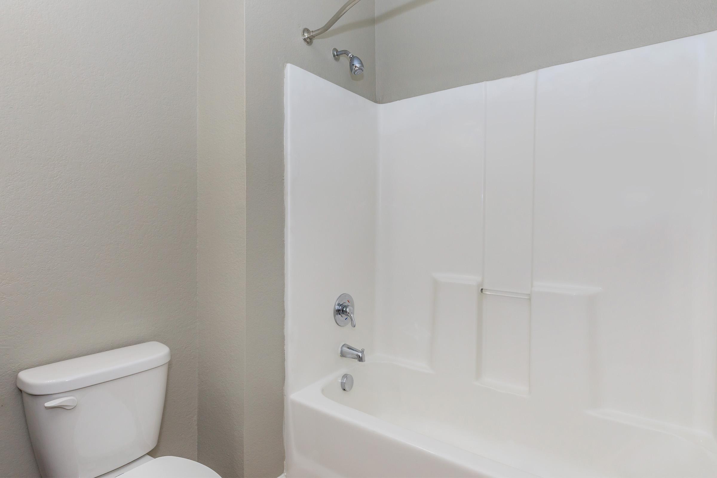 A clean bathroom featuring a white bathtub with a showerhead, a wall-mounted faucet, and a toilet nearby. The walls are painted in a light gray color, creating a modern and minimalistic atmosphere.