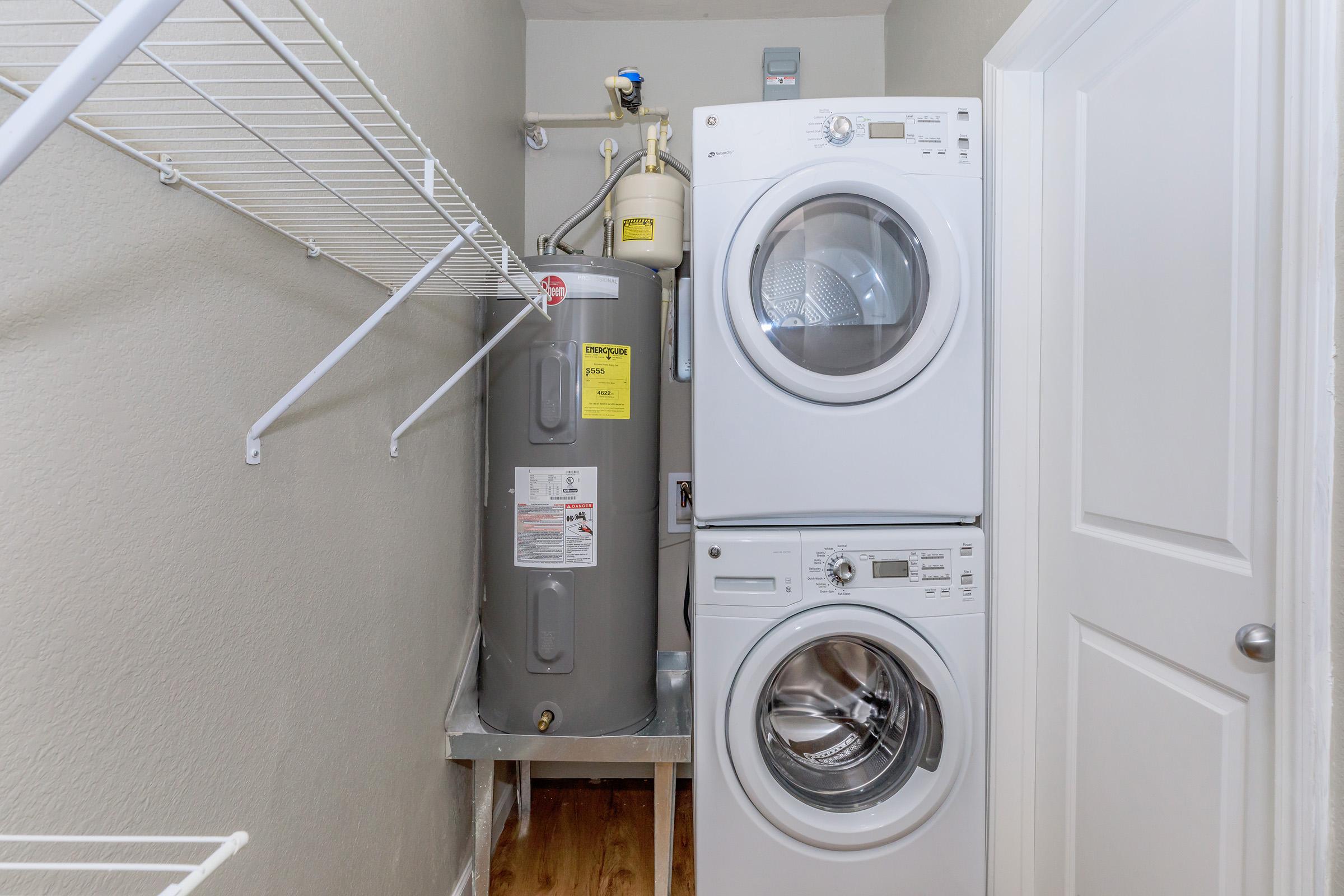 A laundry room featuring a stacked washer and dryer unit next to a water heater. The space is small and includes a wire shelf for storage above the appliances, with light-colored walls and a wooden floor. A closed door is partially visible on the right side.