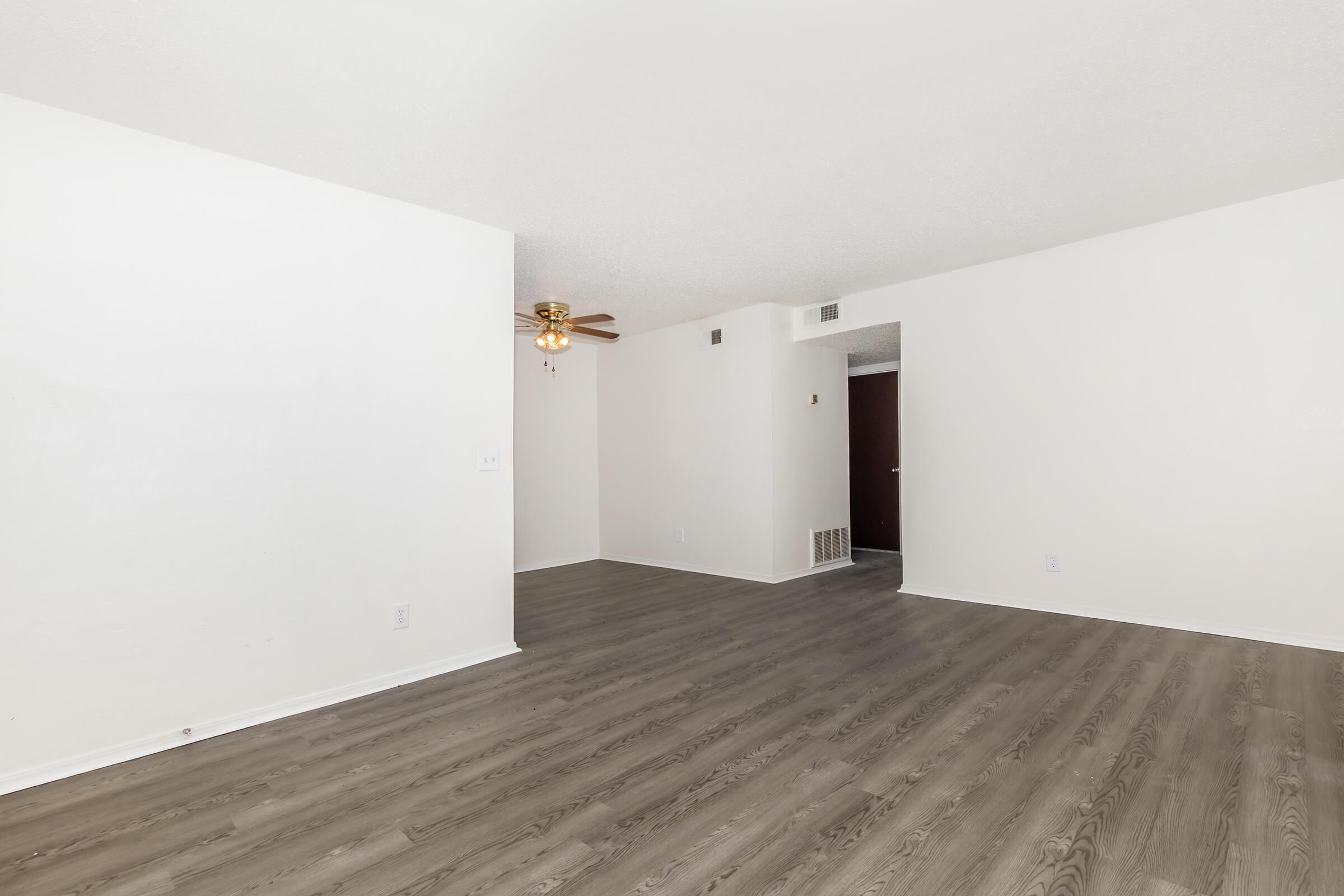 A spacious, empty room with light-colored walls and laminate flooring. A ceiling fan is mounted in the upper left corner. The room appears bright and clean, with a doorway leading to another room visible in the background.