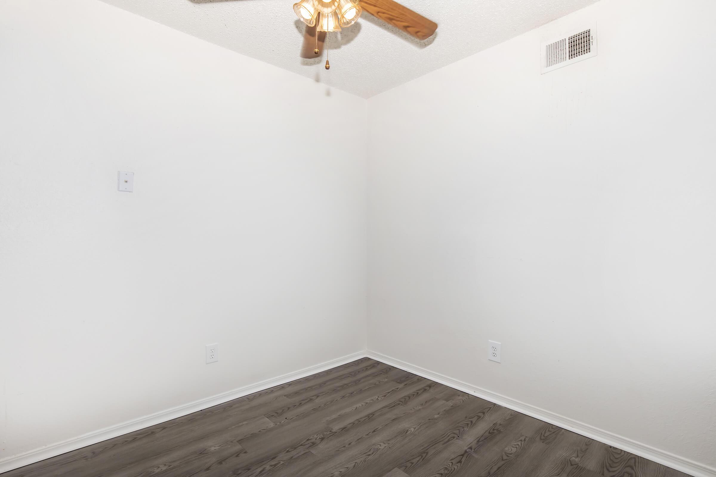 Empty room with light-colored walls and dark wood-style flooring. A ceiling fan with light fixtures is mounted in the corner, and there are two electrical outlets on the wall. The space is well-lit and minimalistic, giving a sense of openness.